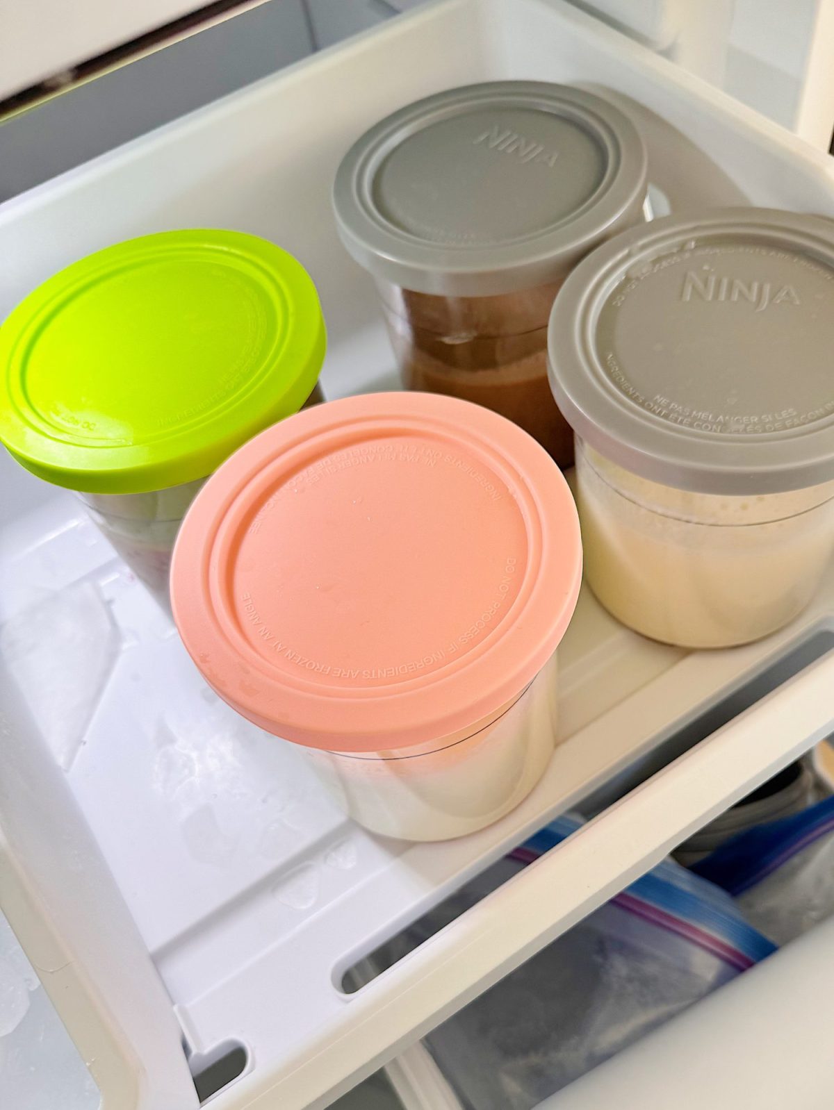 Four plastic containers with colored lids (green, pink, gray, beige) are stored in a refrigerator drawer.
