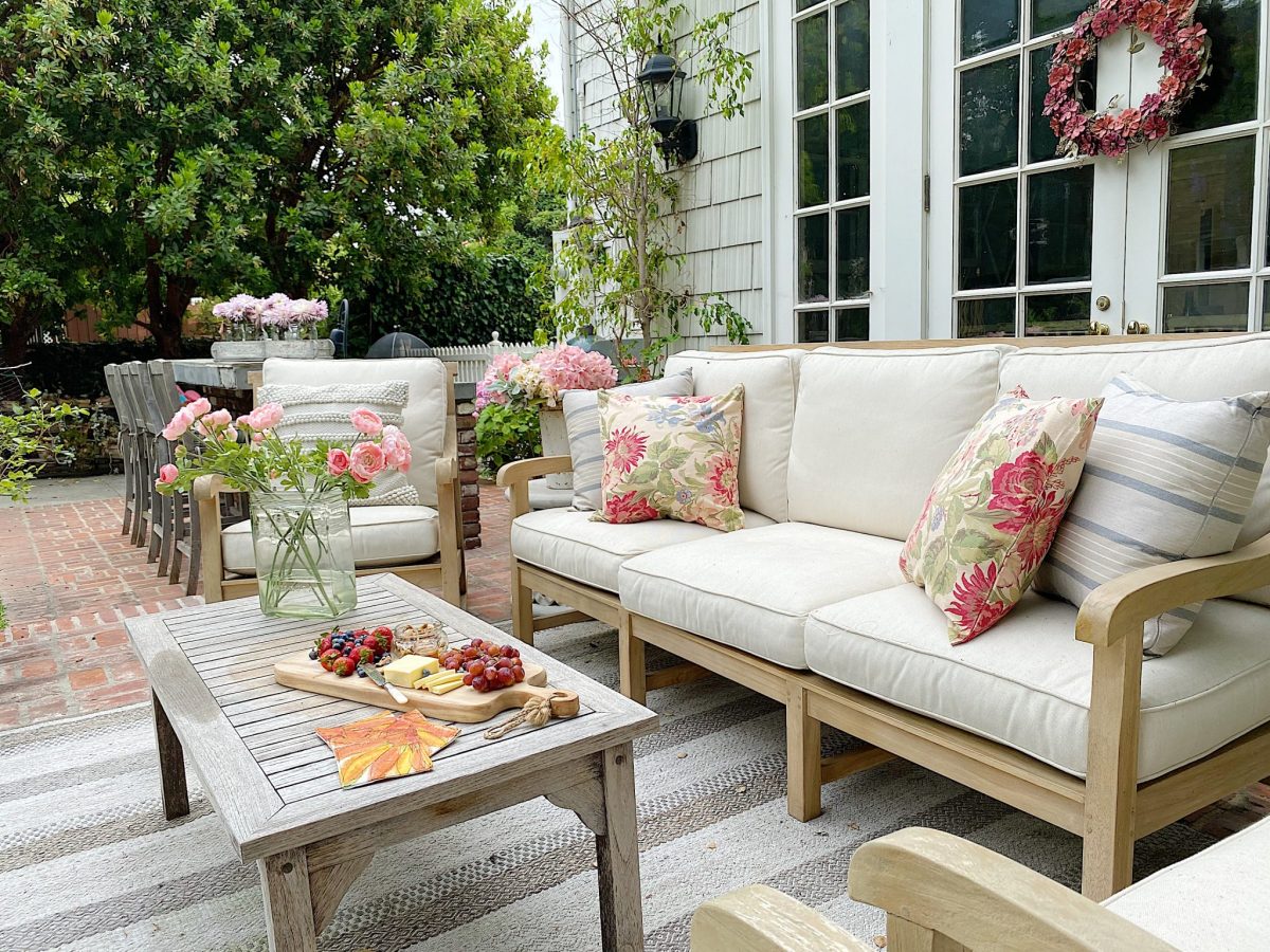 An outdoor patio with cushioned wooden furniture, a vase of pink flowers, a table with cheese and fruit, and a wreath on a door. Greenery and pink flowers surround the area.