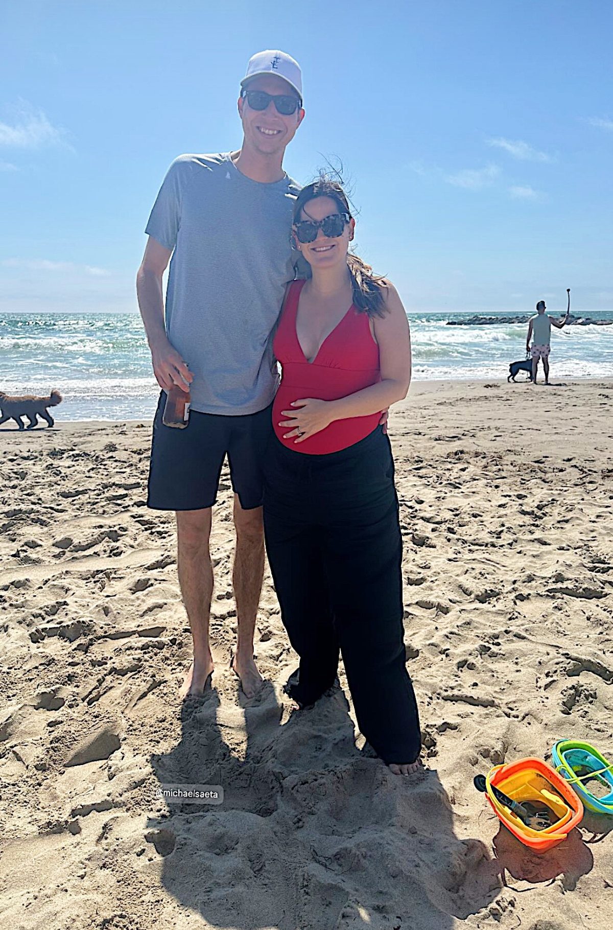 A man and woman stand together on a sandy beach with the ocean in the background. The man holds a drink, and beach toys are visible in the sand. Other people and a dog are in the distance.