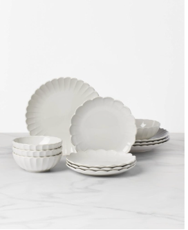 A set of white scalloped-edge plates and bowls is arranged on a marble surface against a white background, perfect for serving guests at your outdoor grilling station.