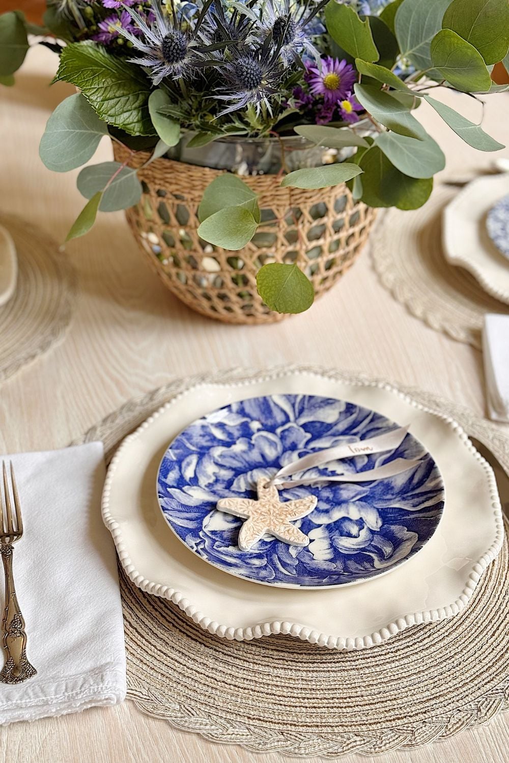 A dining table is set with a beige placemat, white plate, blue floral salad plate, napkin with fork, and a centerpiece with a woven basket holding purple flowers and greenery.