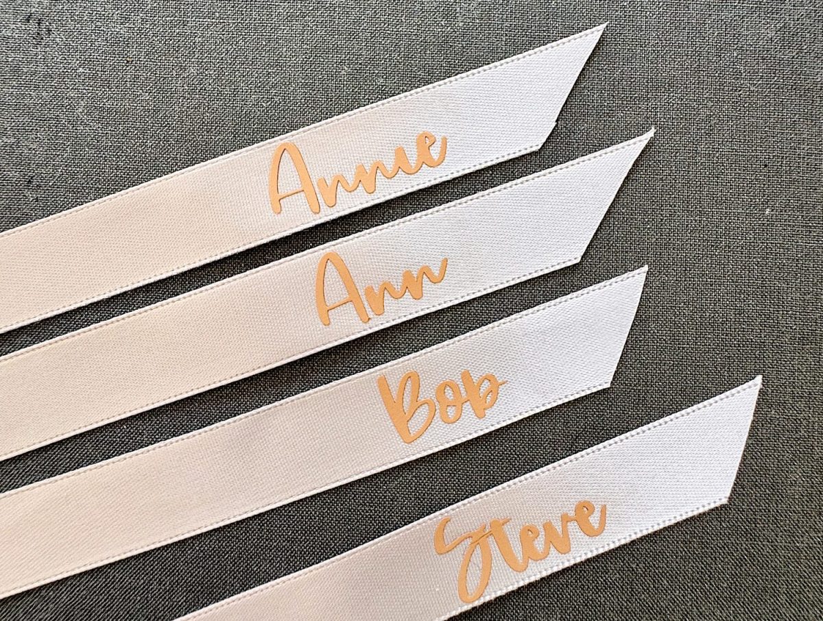 Four white ribbon strips with gold cursive names: Anne, Ann, Bob, and Steve, laid out on a gray fabric background.