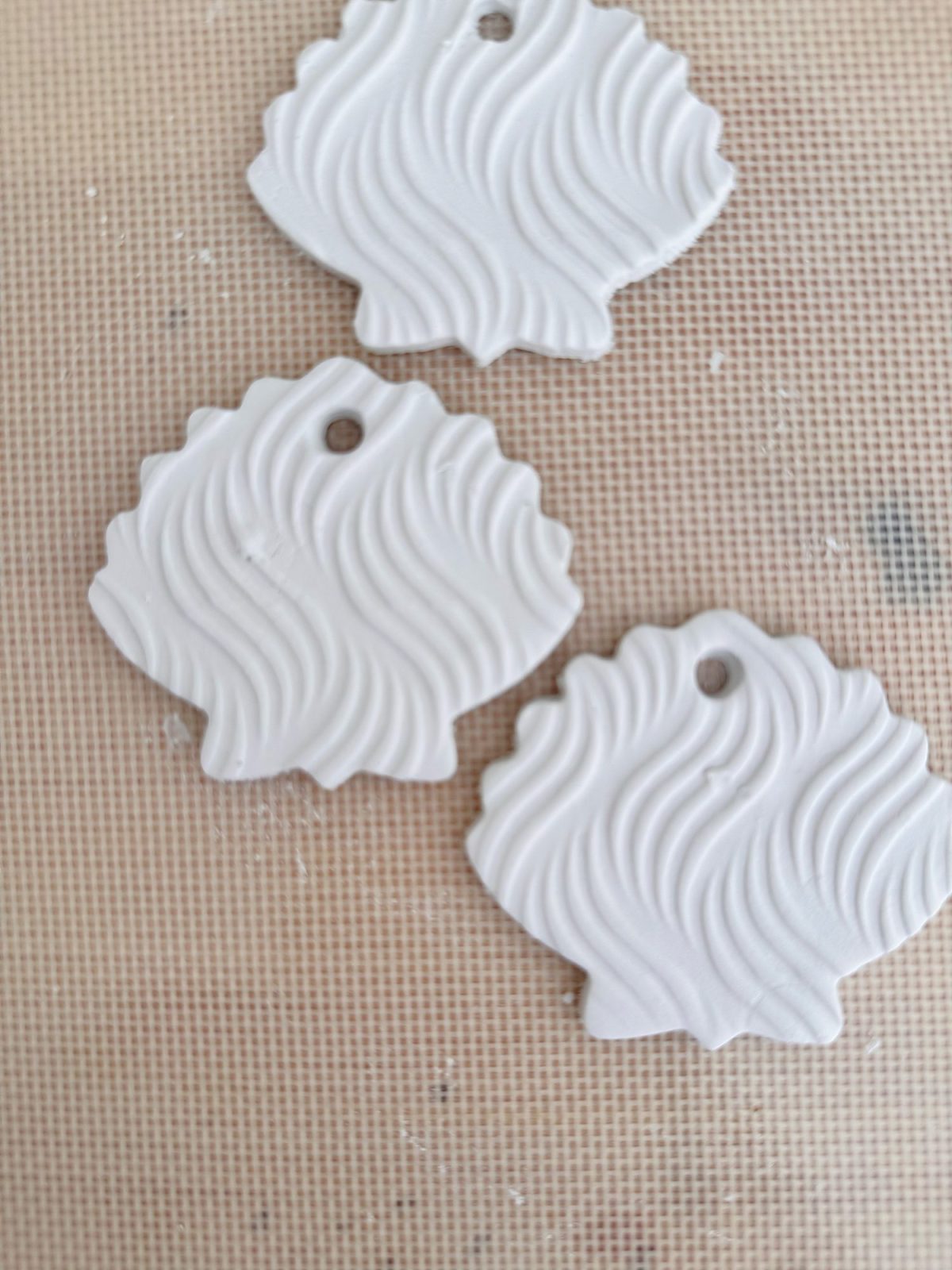 Three white, shell-shaped clay ornaments with wavy textured patterns and small holes at the top, placed on a beige surface.