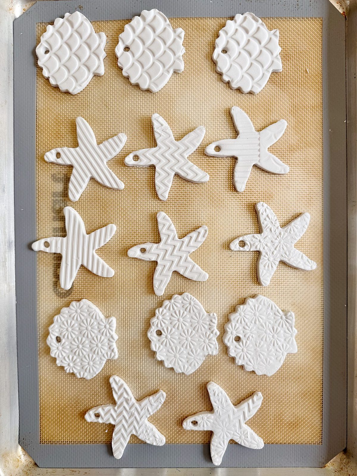 White clay ornaments in shapes of pineapples, starfish, and round medallions with textured patterns are arranged on a silicone mat-lined baking tray.