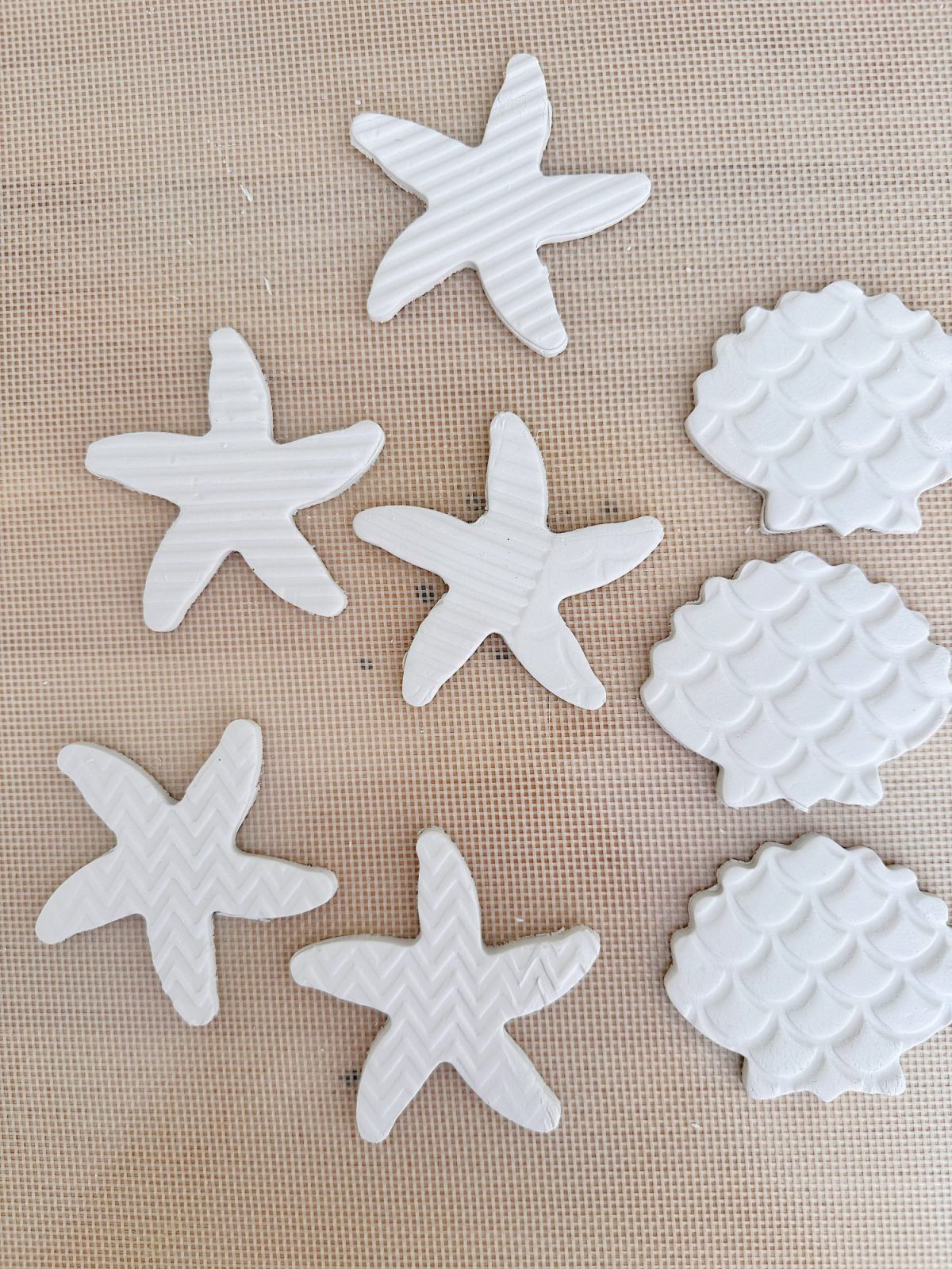 Seven white starfish-shaped and four shell-shaped clay pieces with textured surfaces are arranged on a tan mesh background.