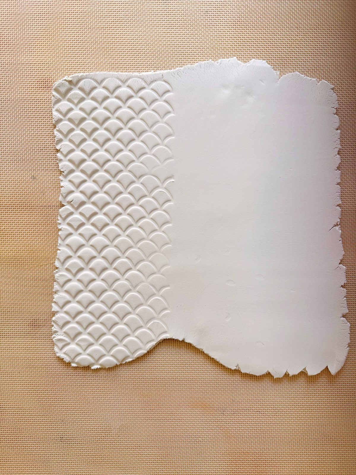 A sheet of white clay with a scalloped pattern on the left half and a smooth texture on the right, placed on a textured tan surface.