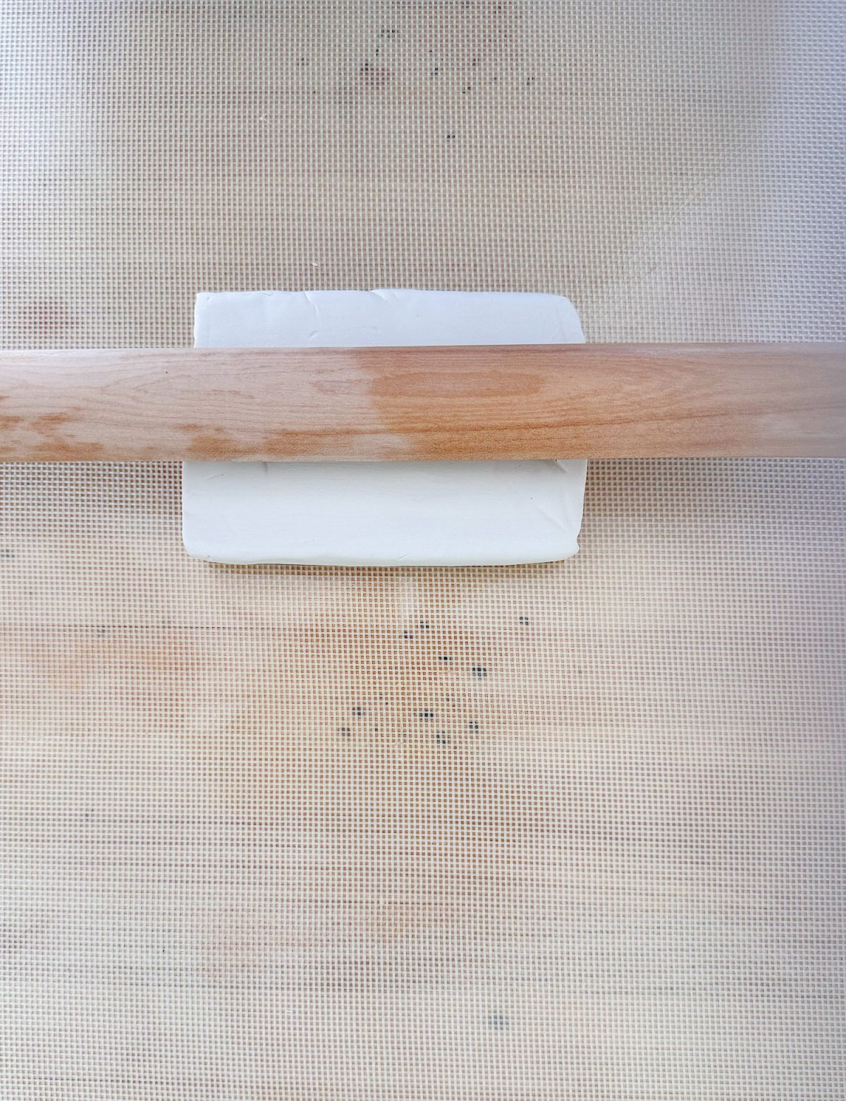 A rolling pin partially flattens a rectangular block of butter on a mesh-patterned surface.