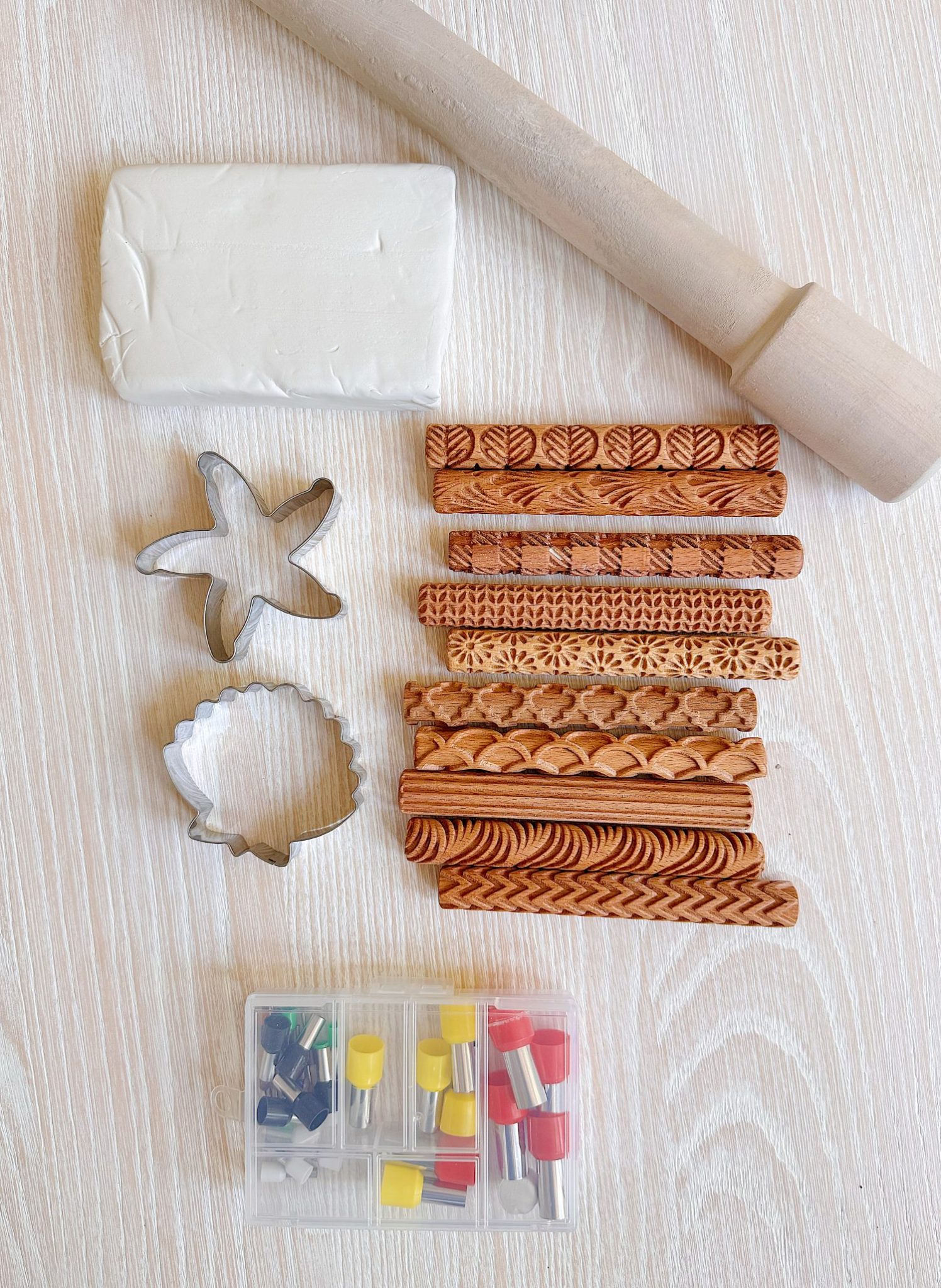 Assorted clay crafting tools on a wooden surface, including a rolling pin, block of white clay, two metal cutters, eight patterned rolling pins, and a box of small tools.