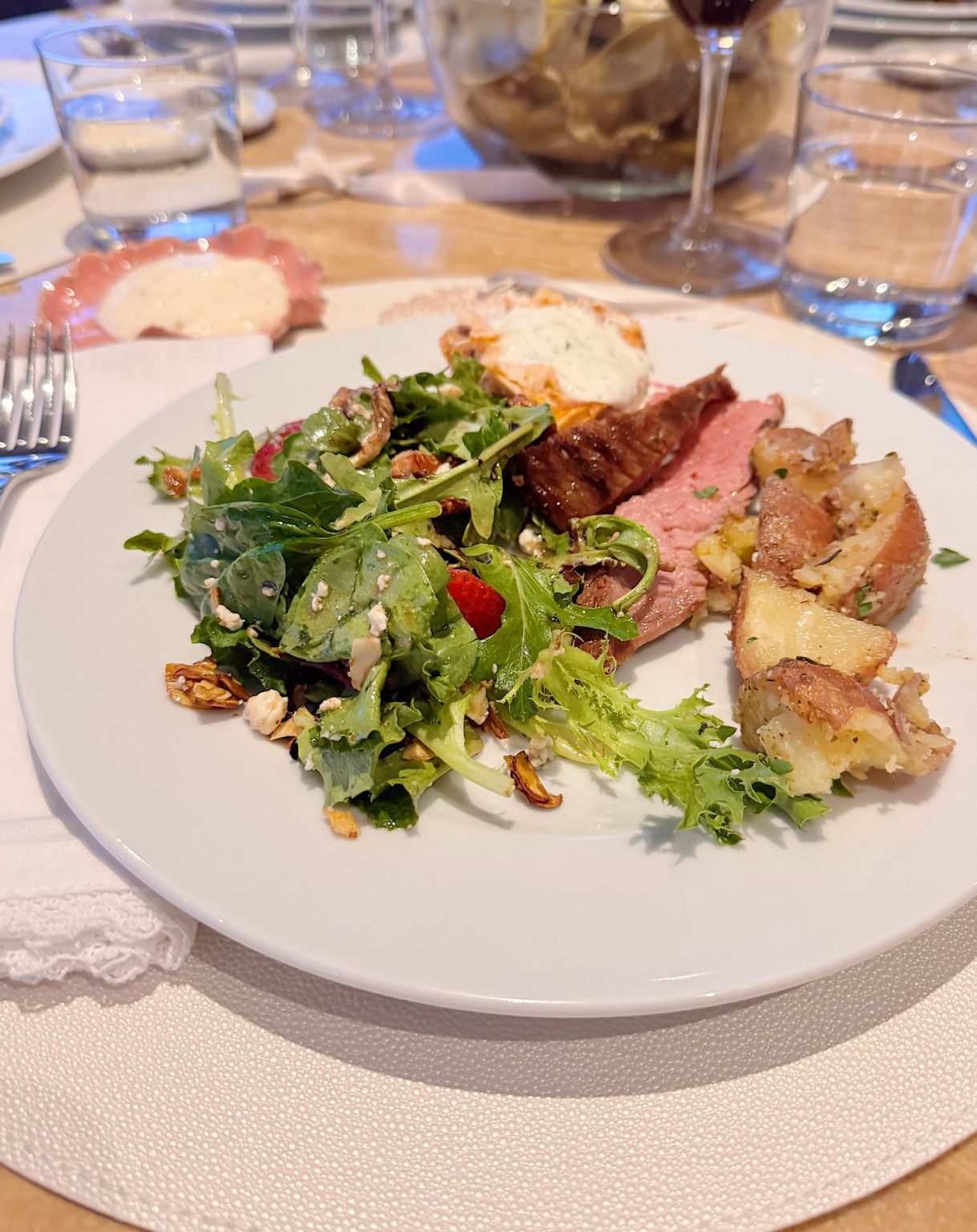 A dinner plate with salad, sliced roast beef, twice-baked potato, crispy potato, and a filled deviled egg, set on a table with glasses and cutlery.