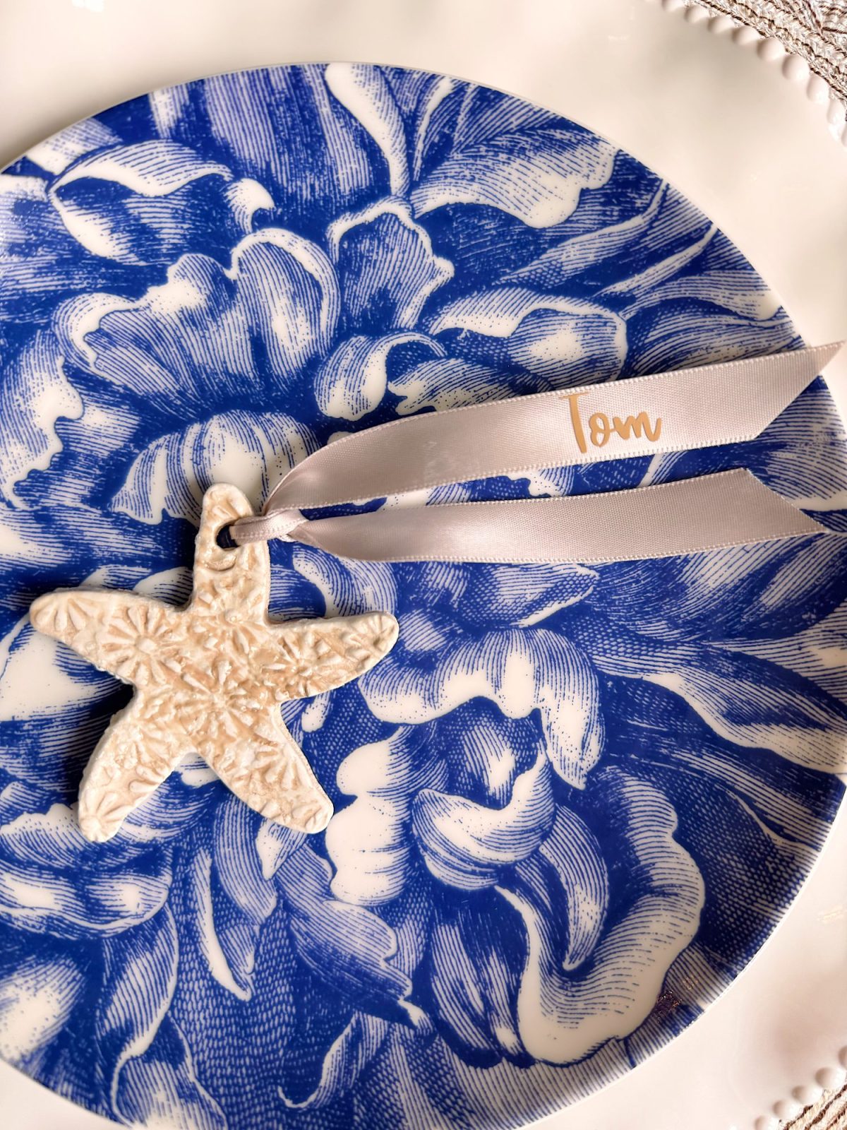 A white ceramic star ornament with a textured pattern and cream ribbon labeled "orn" rests on a blue and white floral plate.
