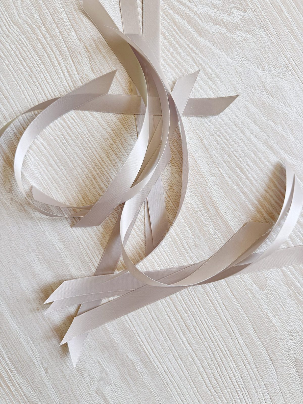 Several pale beige satin ribbons with pointed ends are loosely arranged on a light wood surface.