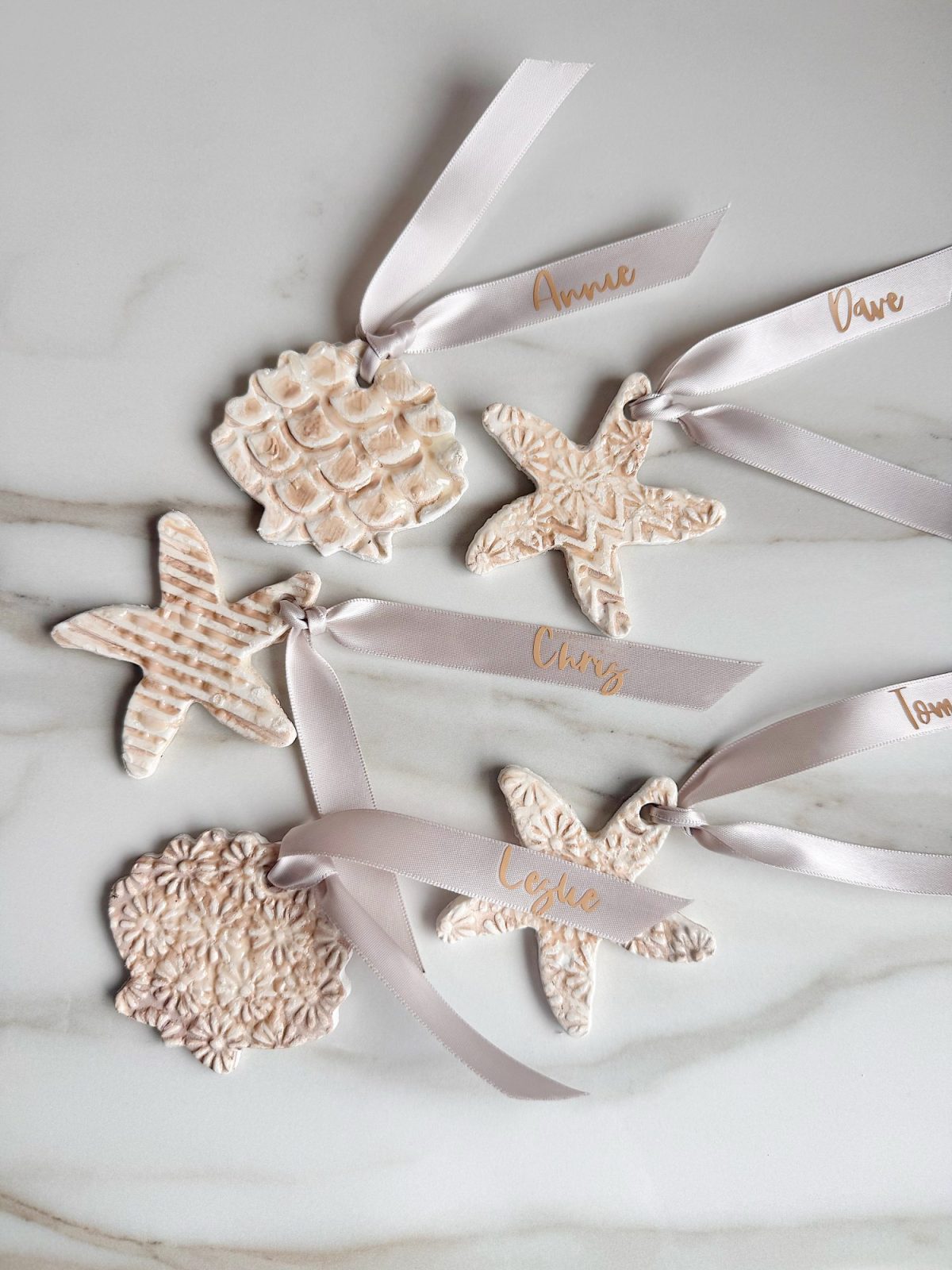 Five textured ceramic ornaments with pale ribbons, shaped like starfish and shells, each labeled with a different name in gold script, laid out on a marble surface.