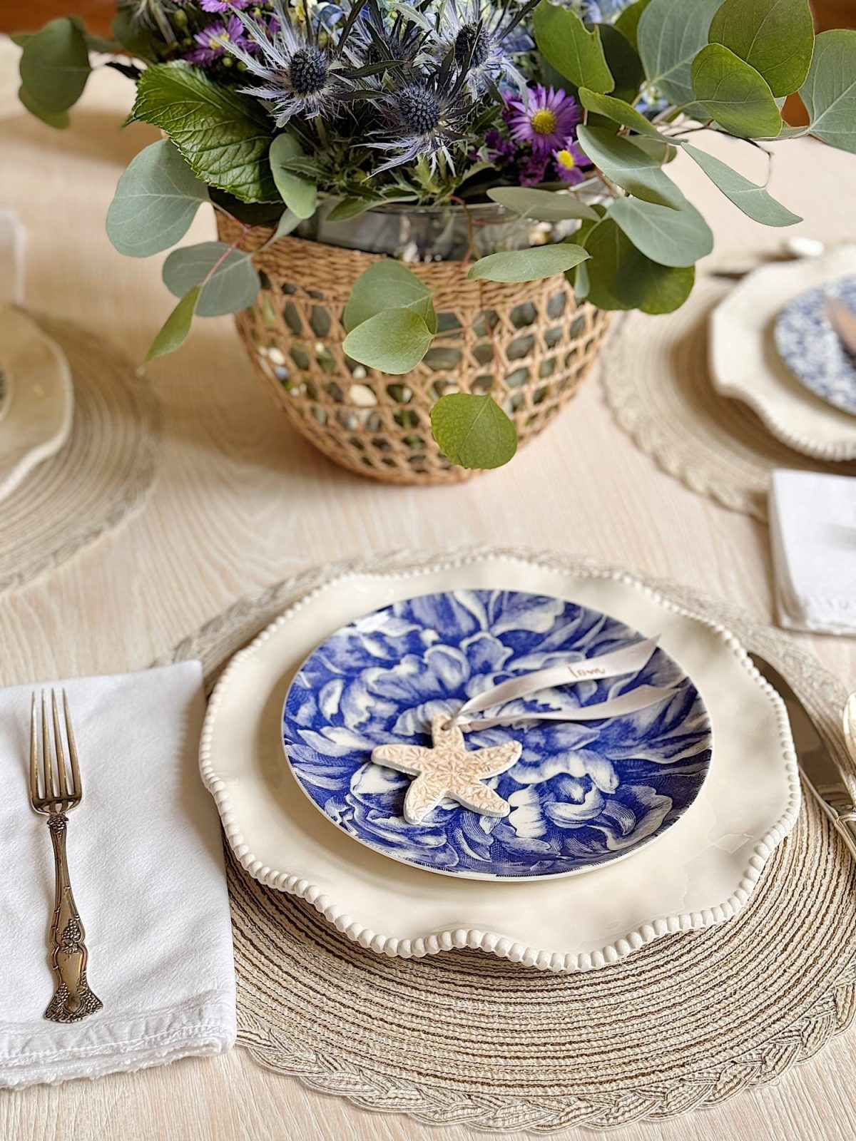 A table set with a blue and white floral plate on a cream charger, a fork on a white napkin, and a vase with purple flowers and greenery as the centerpiece.