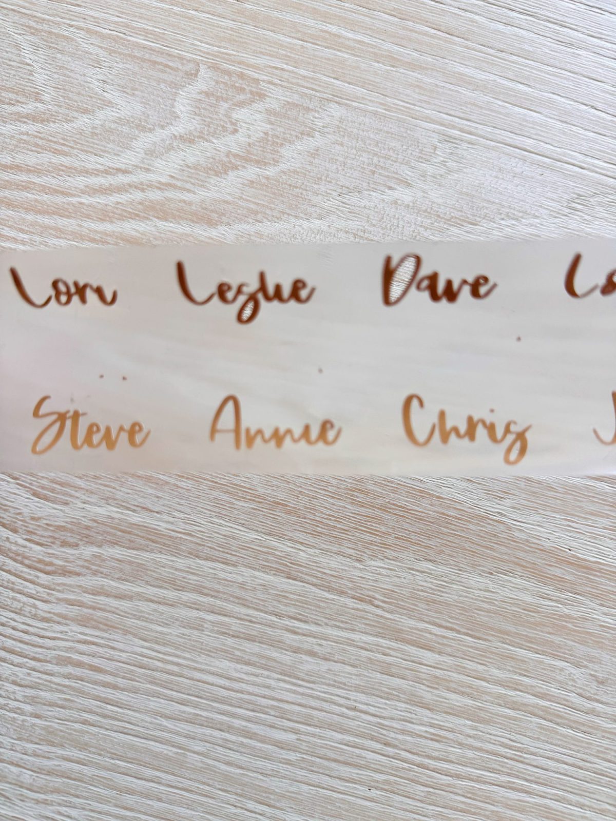 A sheet of transparent stickers displays names in cursive, including Lori, Leslie, Dave, Steve, Annie, and Chris, on a light wood surface.