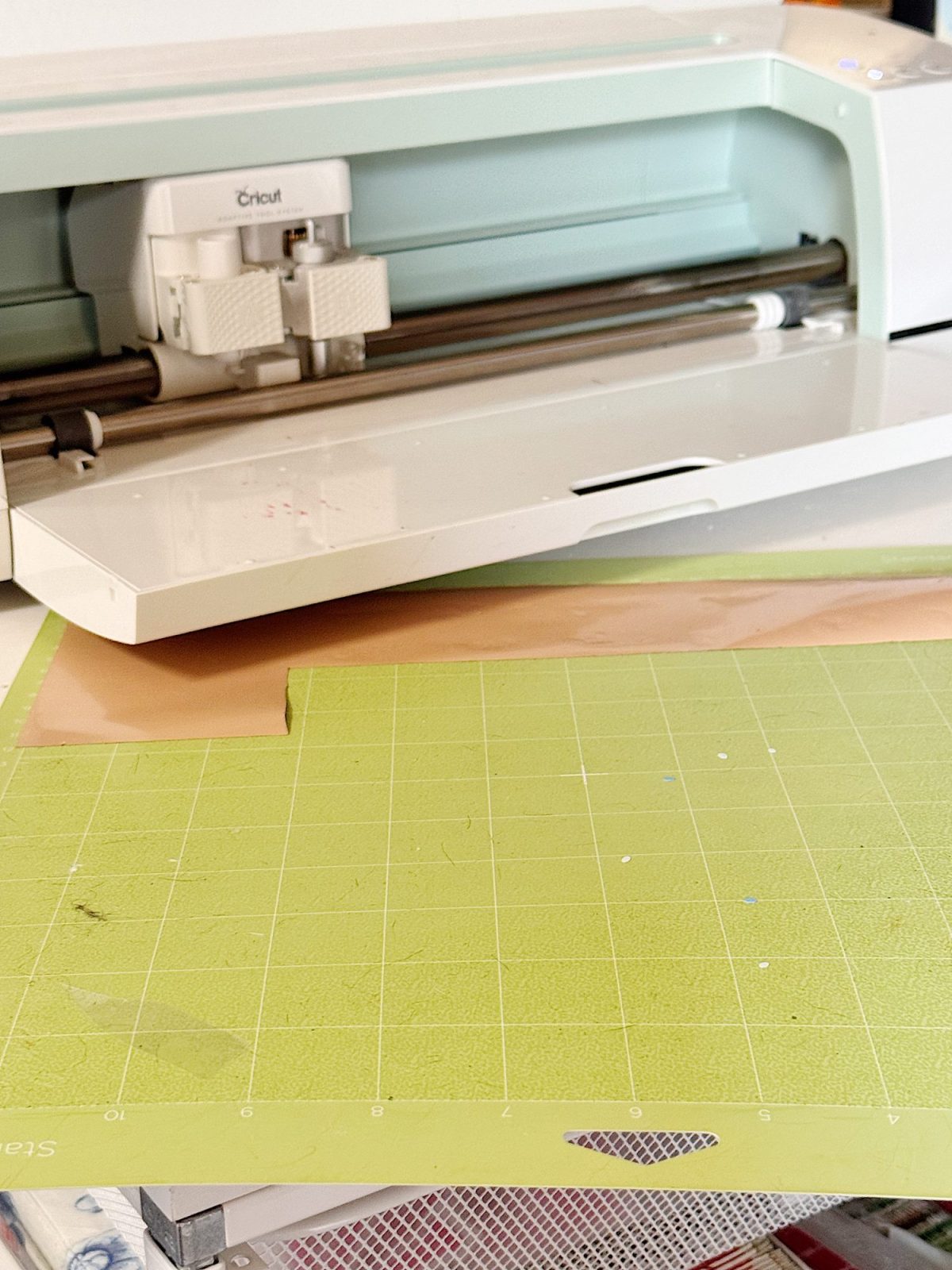 A Cricut cutting machine with its lid open, a piece of brown material loaded inside, and a green cutting mat positioned in front.