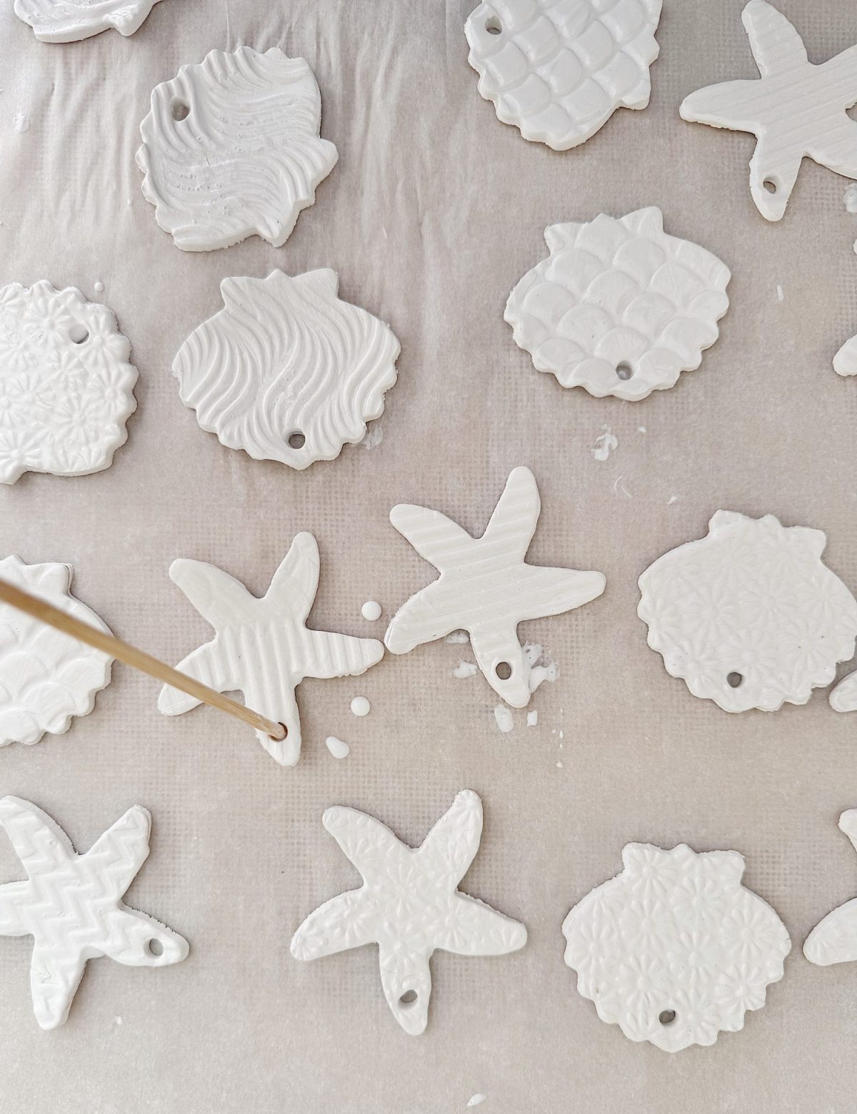 White clay ornaments in various shapes and textured patterns are laid out on a surface, with a hand using a stick to poke a hole in one of the star-shaped pieces.