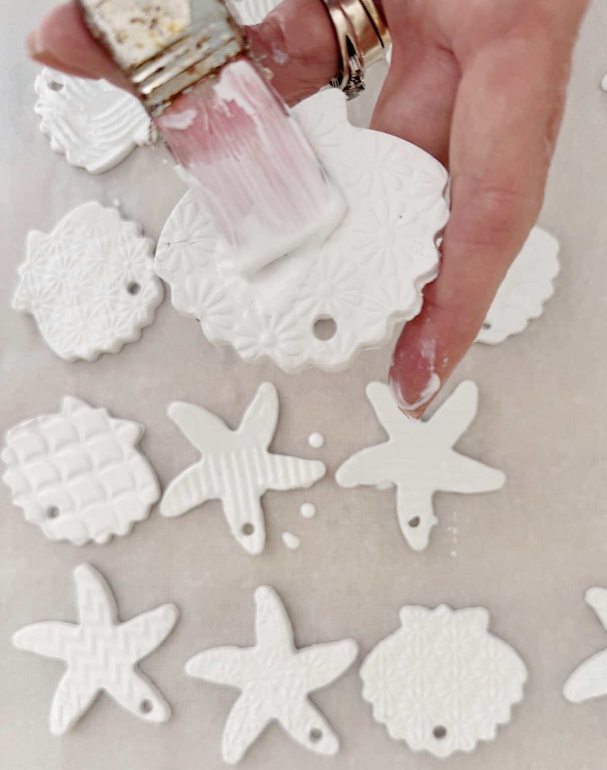 A hand uses a paintbrush to apply white paint to a textured, round ornament among several white starfish and circular clay ornaments on a flat surface.