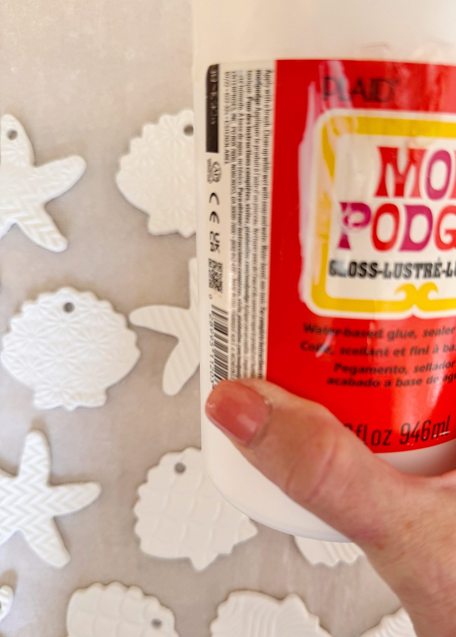 A hand holds a Mod Podge bottle in front of a wall with several white, textured, fish-shaped ornaments.