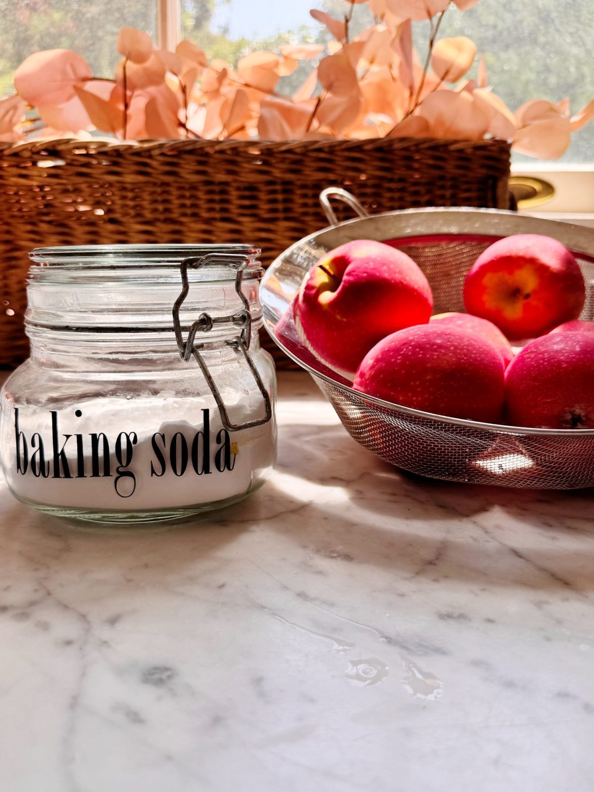 A glass jar labeled "baking soda" sits on a marble countertop next to a metal colander filled with red apples; a wicker basket with dried leaves is in the background.