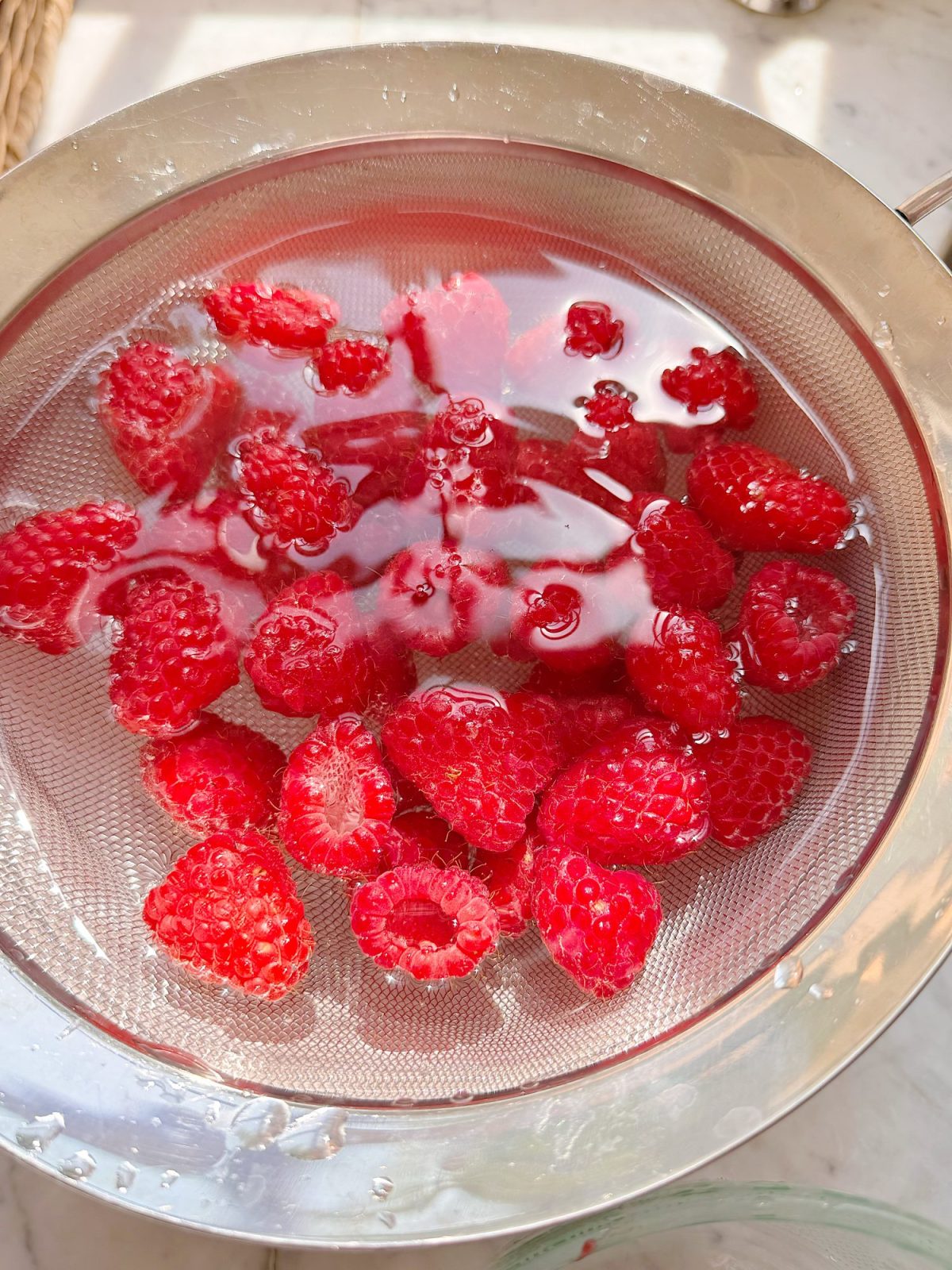 Fresh raspberries soaking in water inside a metal mesh strainer, placed over a bowl on a light surface.