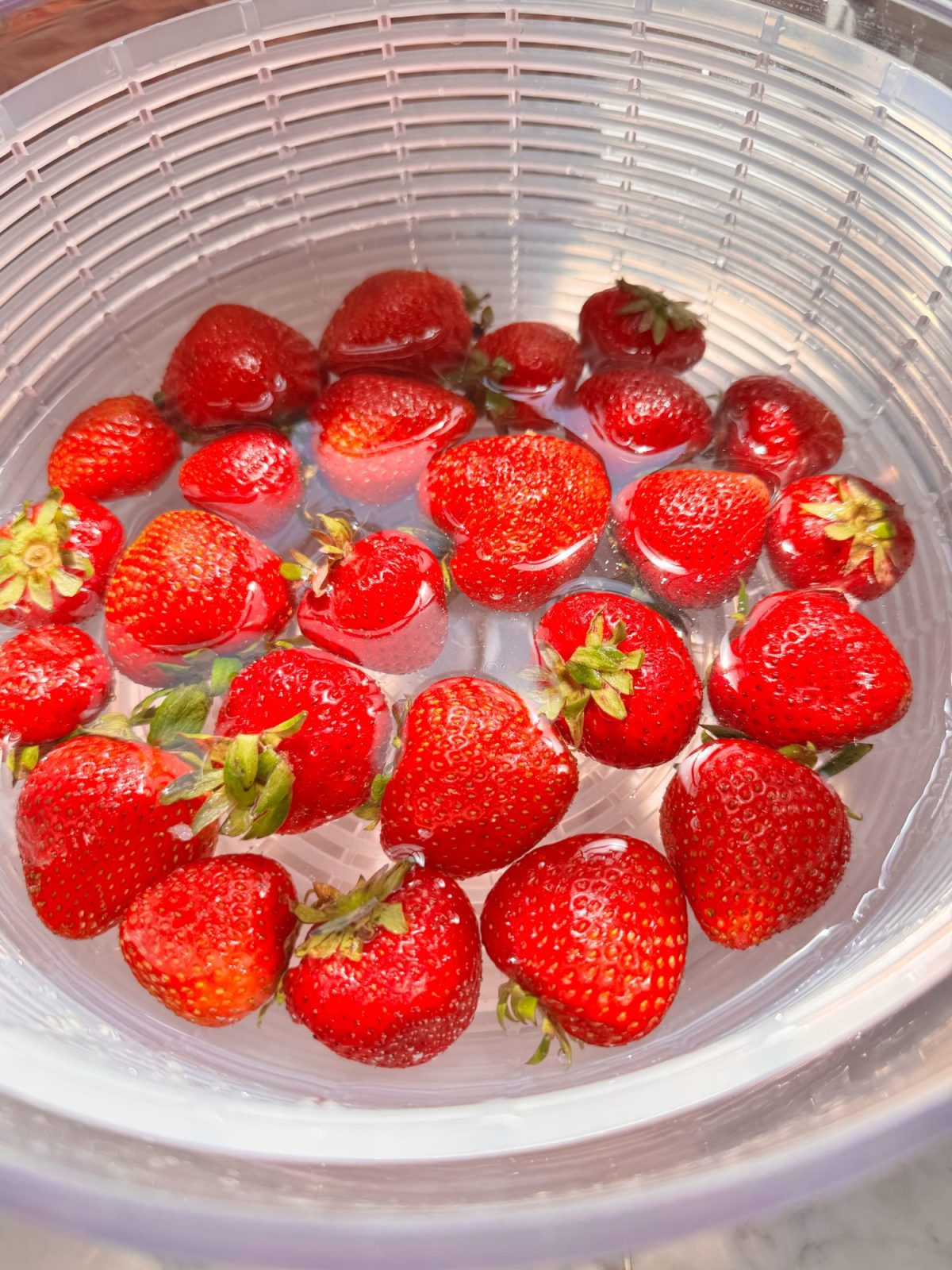 Fresh strawberries soaking in a clear plastic bowl filled with water.