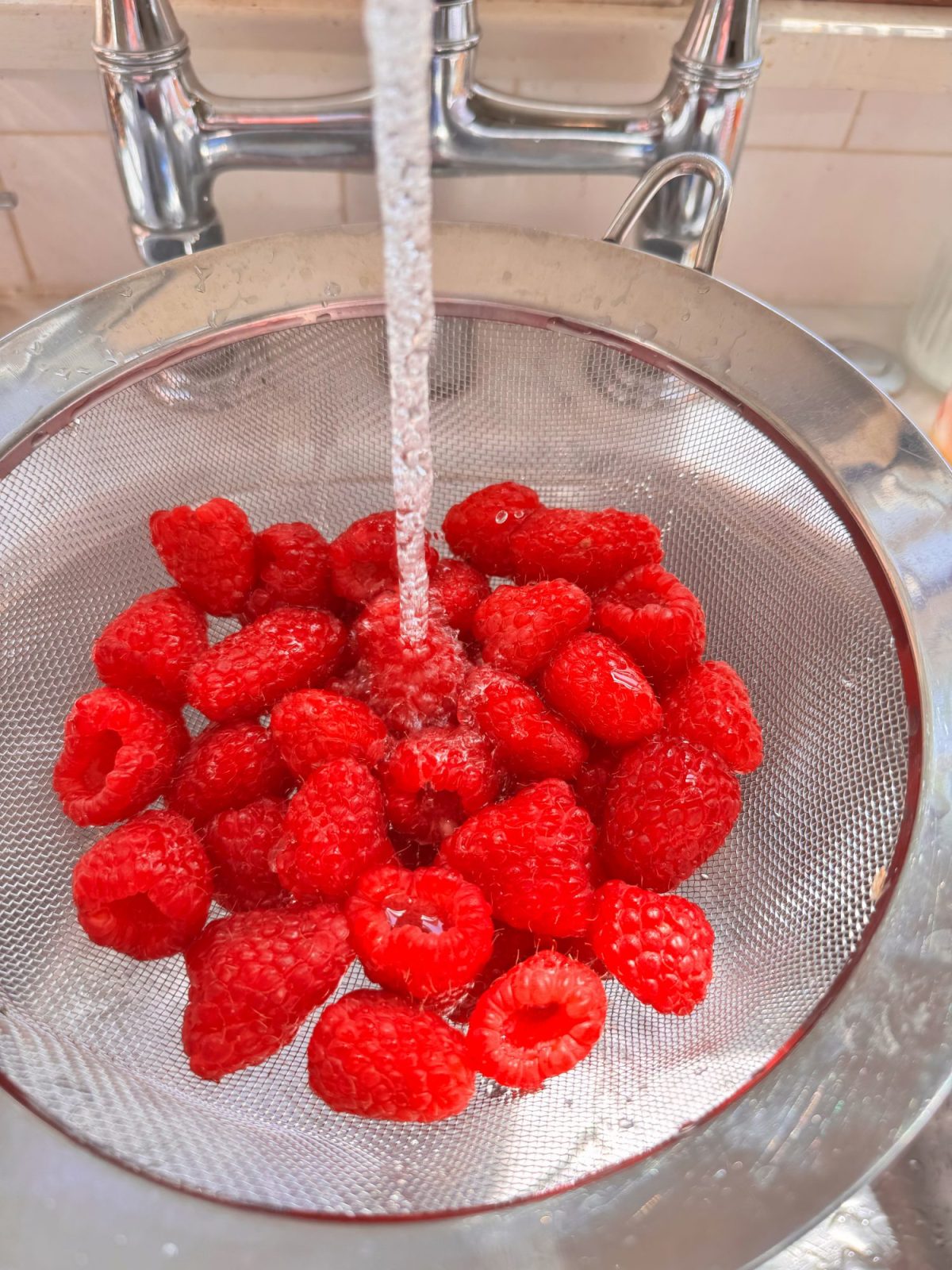 Fresh raspberries being rinsed under running water in a metal mesh strainer in a kitchen sink.