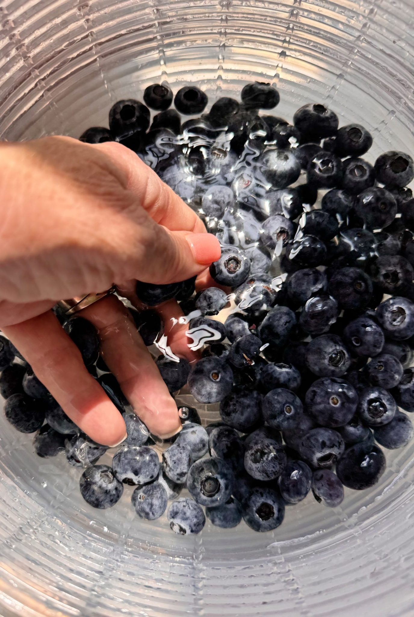 A hand rinses fresh blueberries in a clear glass bowl filled with water.