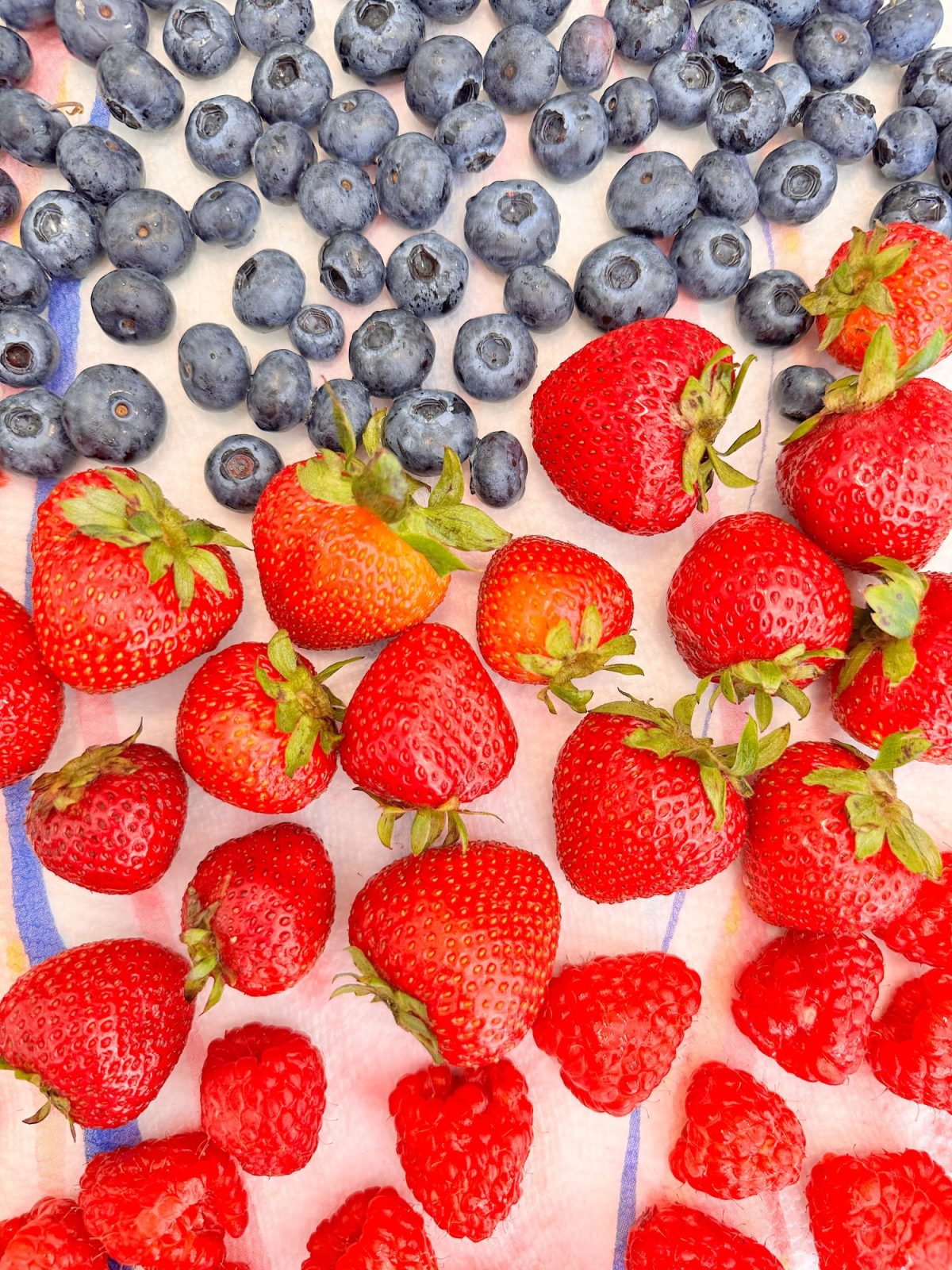Blueberries, strawberries, and raspberries arranged in rows on a white surface with blue and red stripes, resembling the American flag.