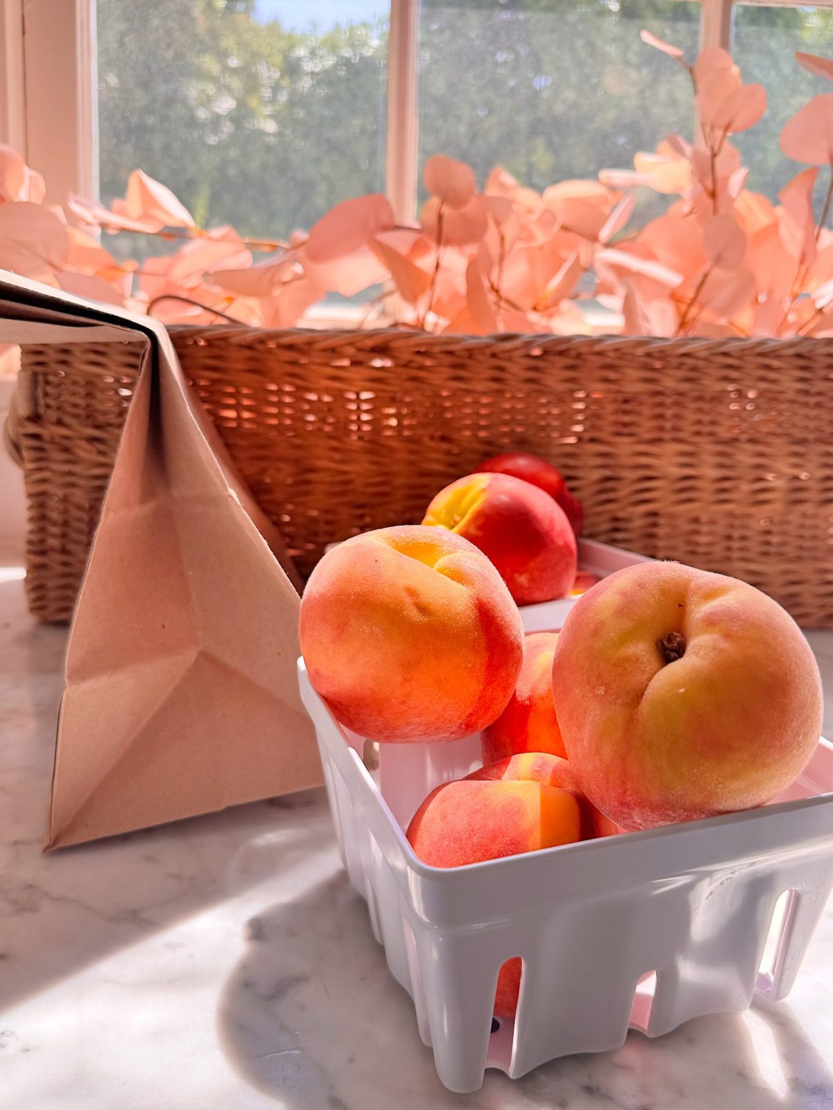 A white basket of peaches sits on a marble countertop near a brown basket and dried pink leaves, with sunlight streaming through a window.