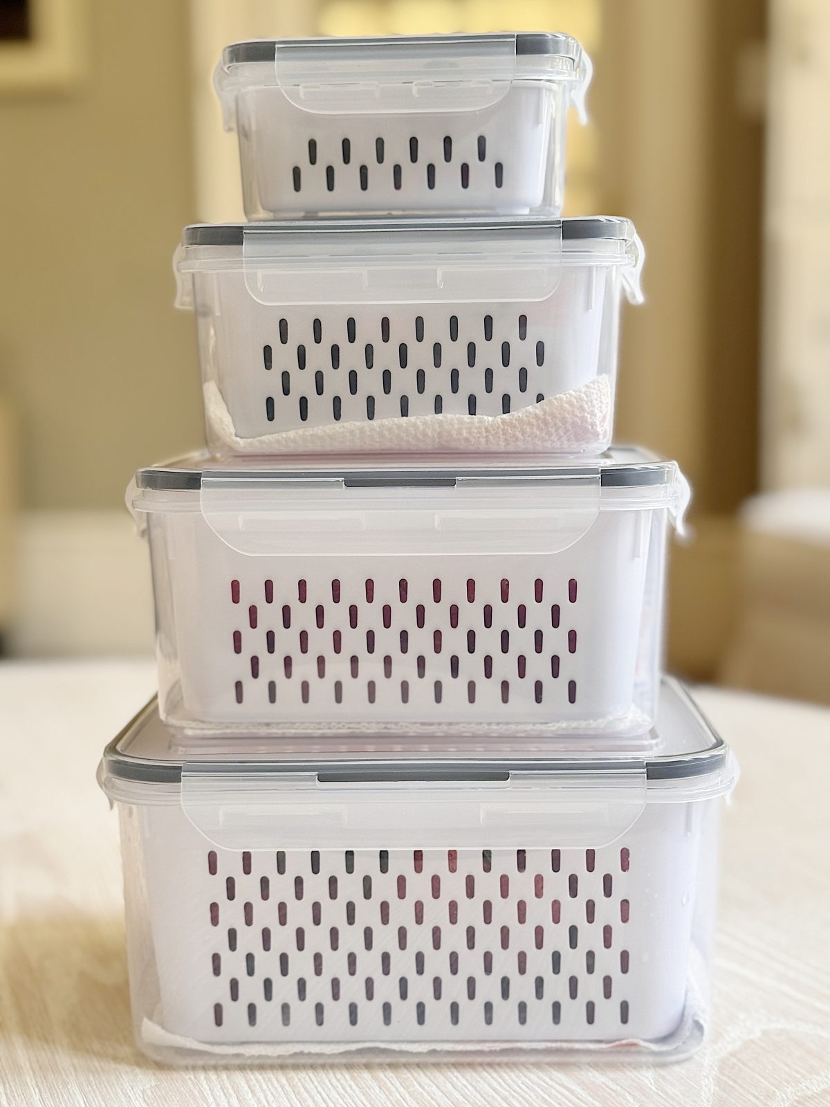 Four transparent plastic storage containers with vented lids are stacked on top of each other on a light-colored surface.