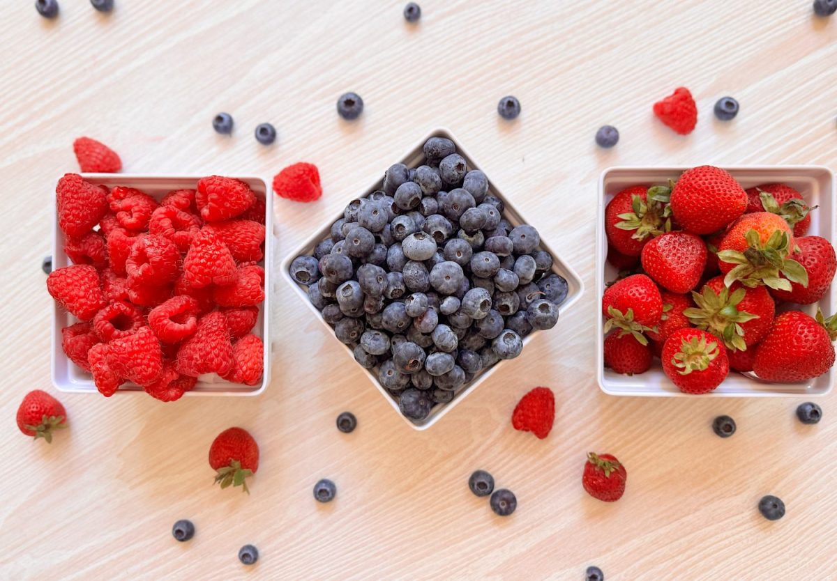 Three square bowls filled with raspberries, blueberries, and strawberries are arranged on a light wooden surface with some loose berries scattered around.