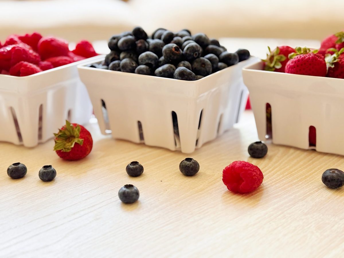 Three white containers filled with raspberries, blueberries, and strawberries sit on a light wooden surface, with some berries scattered around them.