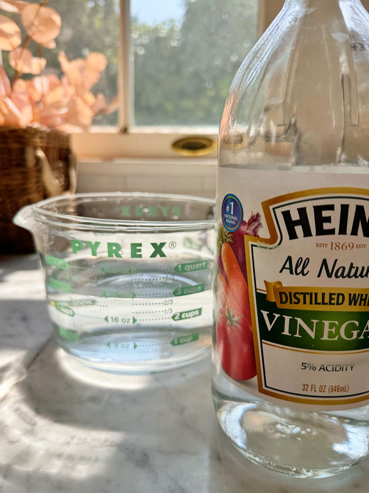 A clear Pyrex measuring cup with water sits on a marble countertop next to a bottle of Heinz distilled white vinegar. Sunlight streams through a window in the background.