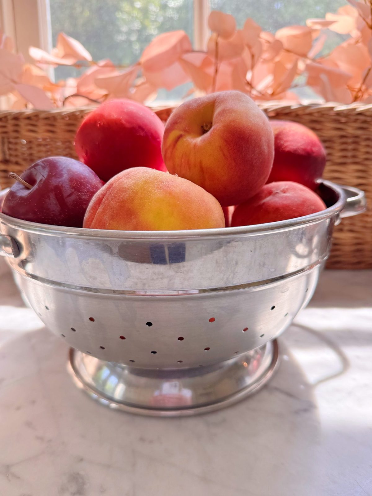 A metal colander filled with peaches and nectarines sits on a marble countertop, with sunlight shining through a window and dried leaves in the background.