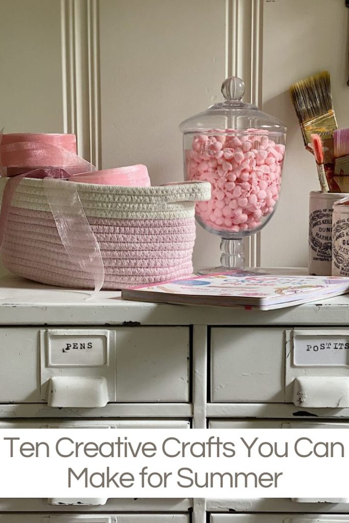 A pink and white woven basket, ribbon, jar of pink craft supplies, paintbrushes, and a notebook sit on a white drawer unit labeled "PENS" and "POST-ITS." Text reads: "Ten Creative Crafts You Can Make for Summer.