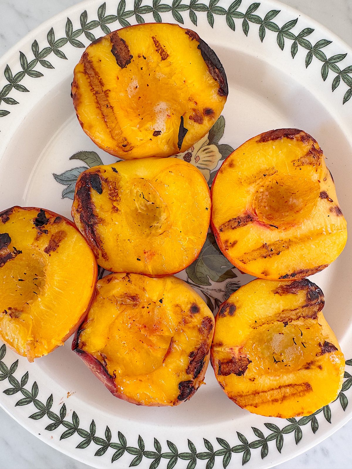 Six grilled peach halves arranged on a decorative white plate with a leaf pattern border.