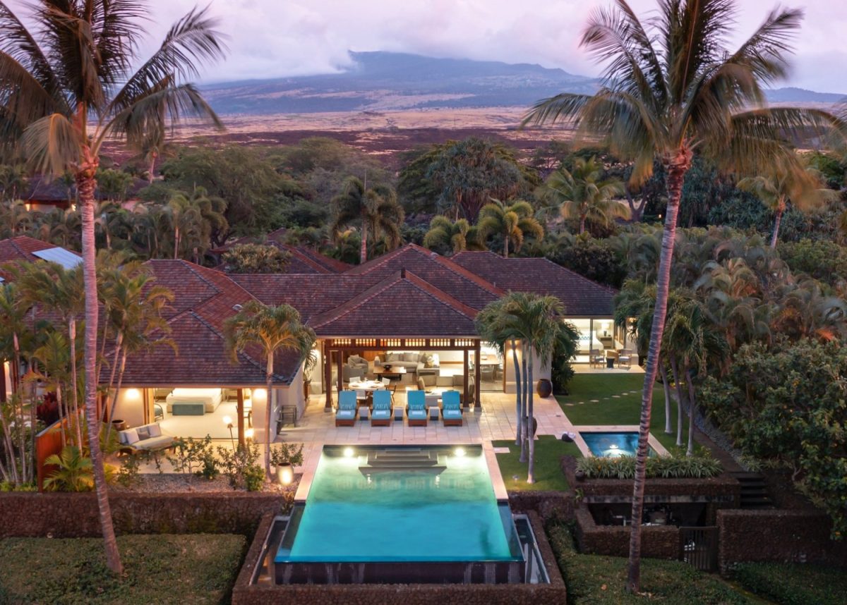 A luxury villa with an infinity pool, outdoor lounge chairs, and palm trees, set against a backdrop of mountains and lush greenery at dusk.