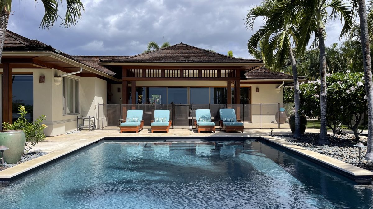 A swimming pool with four blue lounge chairs lined up on the pool deck in front of a modern house with large windows and tropical landscaping.