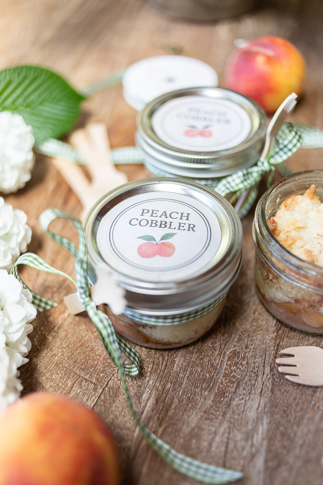 Two glass jars labeled "Peach Cobbler" with ribbon ties are placed on a wooden surface, surrounded by peaches, flowers, and small wooden utensils.