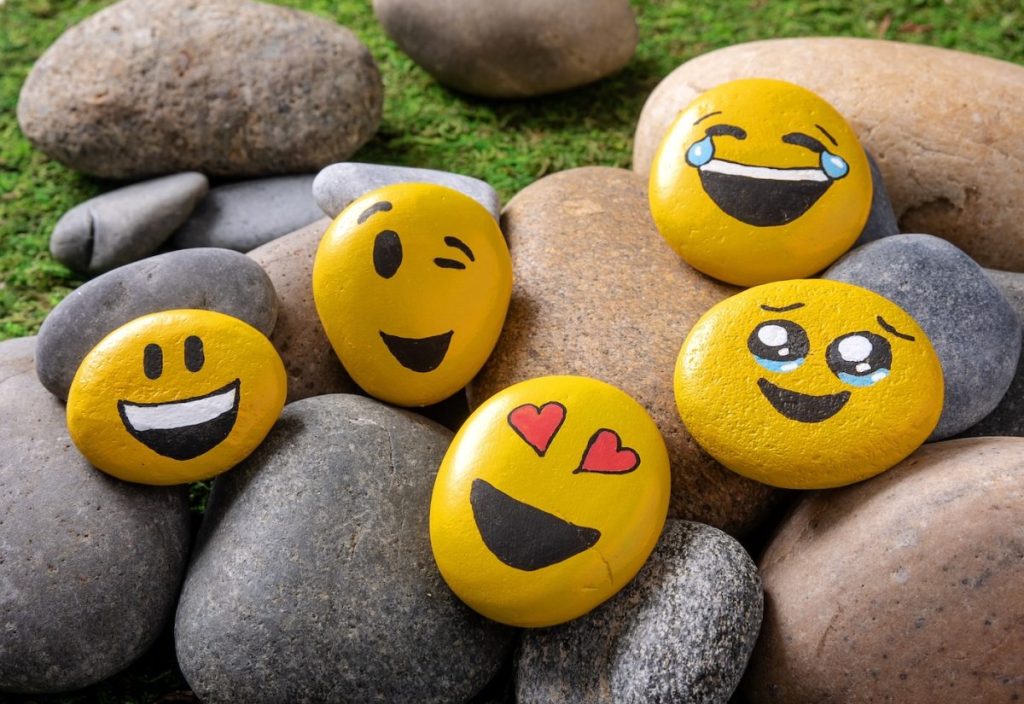 Several smooth stones are painted yellow with various emoji faces, including smiling, winking, laughing, crying, and heart eyes, placed among unpainted rocks on grass.