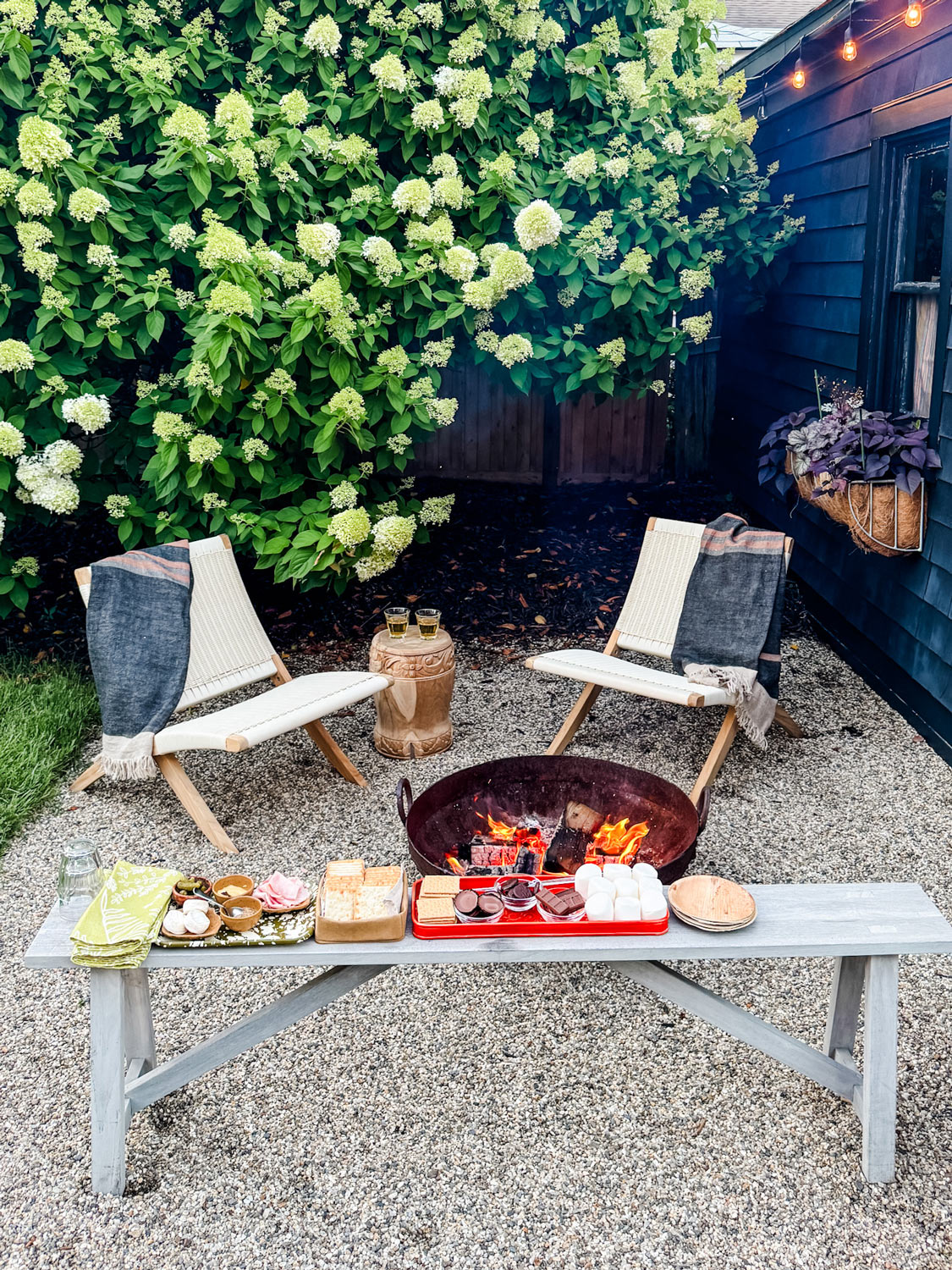 Two chairs and a small table sit by a fire pit, with a bench in front displaying s'mores ingredients, in a gravel backyard with hydrangeas and string lights.