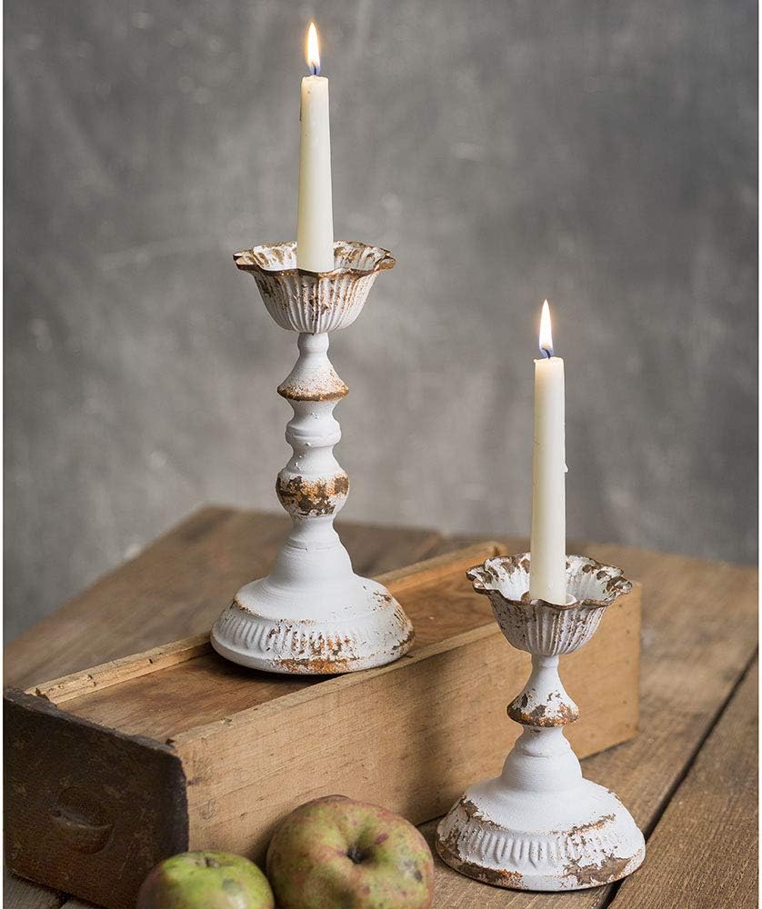 Two lit white candles in distressed white candlesticks sit on a wooden surface next to two apples, creating a cozy scene that says fall is here, all set against a plain gray background.