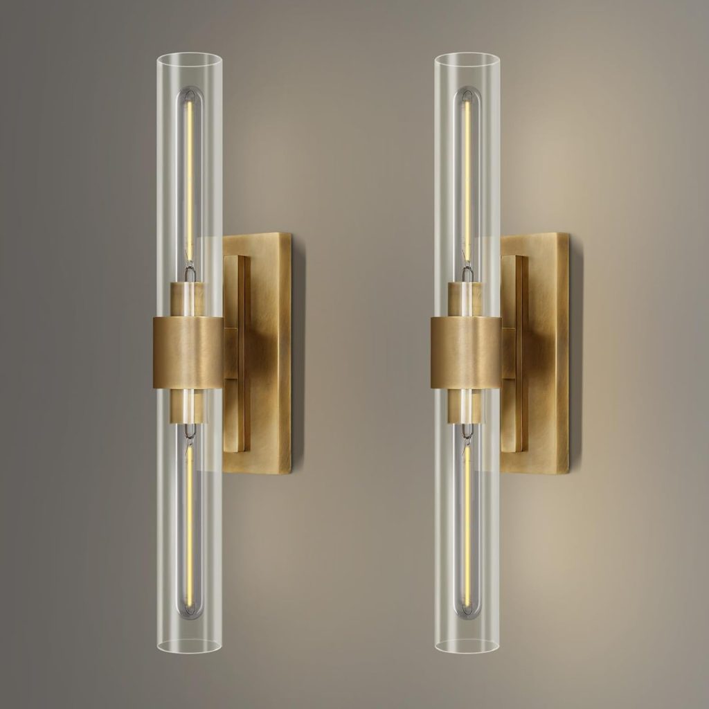 Two modern wall sconces with cylindrical glass shades and brass-finished rectangular bases are mounted on a gray wall.
