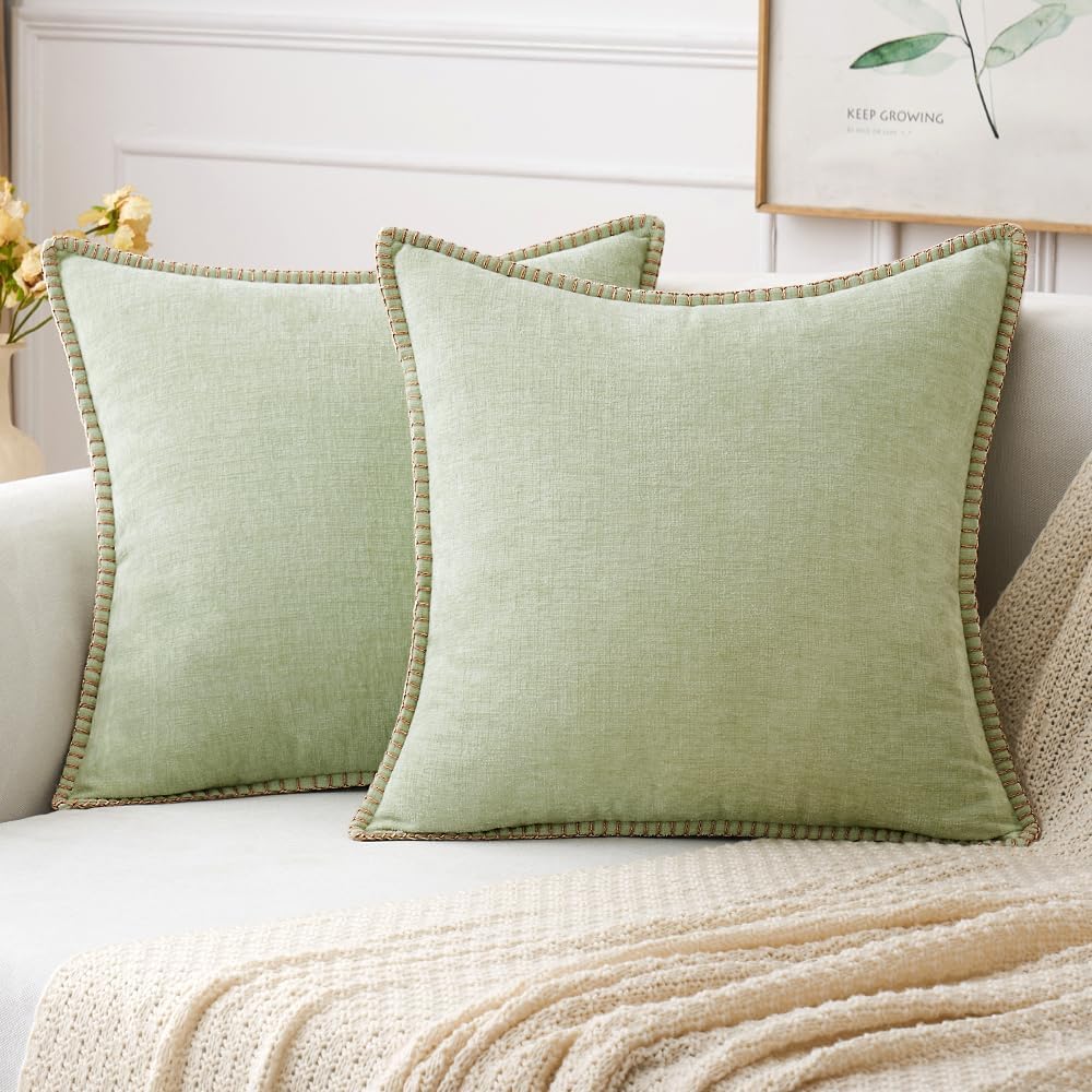 Two light green square pillows with textured edges on a light-colored sofa, next to a beige knitted throw blanket, against a white paneled wall.