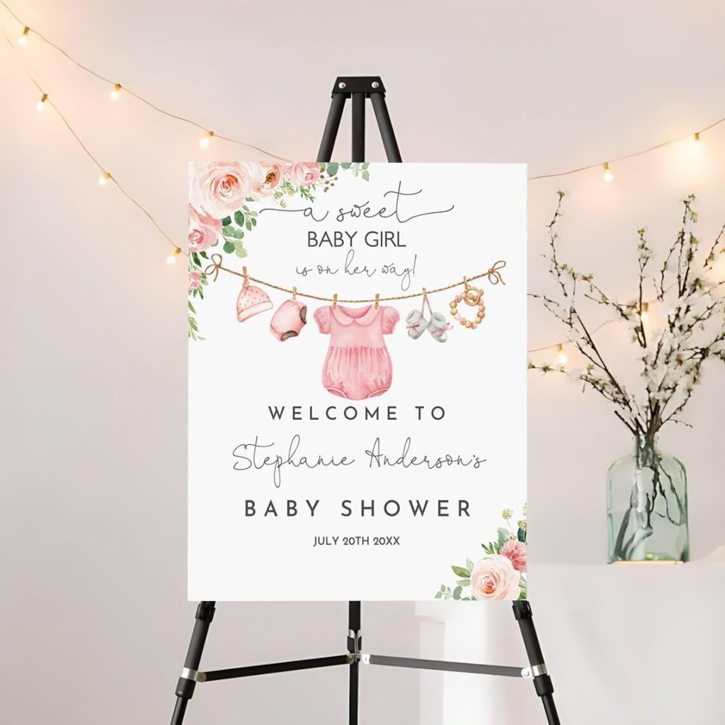 A baby shower welcome sign on an easel reads "A sweet baby girl is on her way! Welcome to Stephanie Anderson's Baby Shower, July 20th 20XX," with floral and baby clothes decorations.