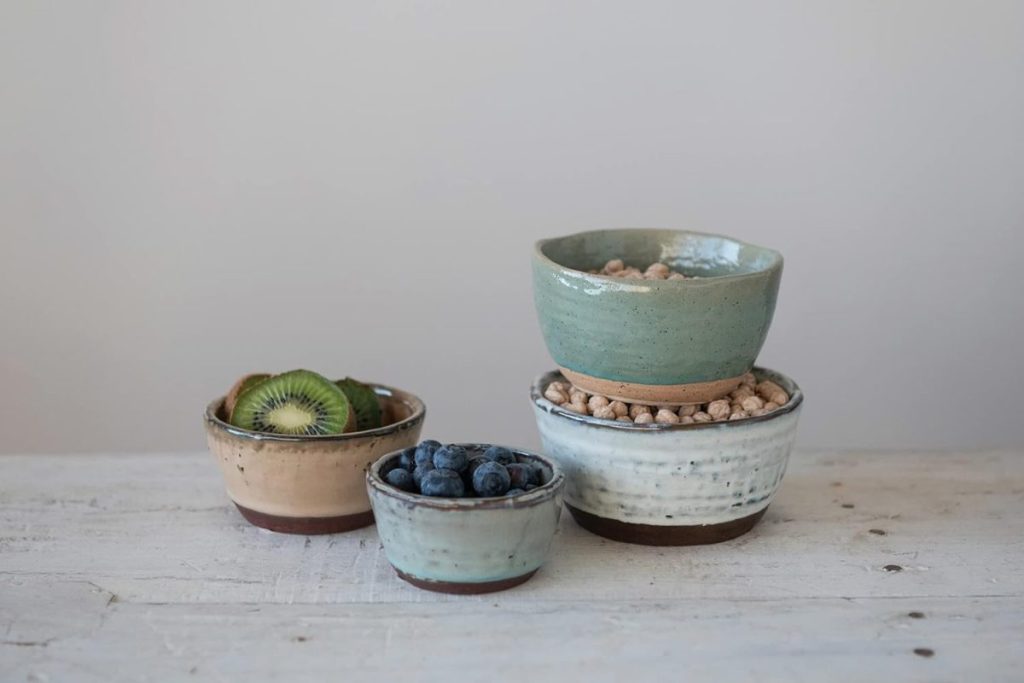 Four handmade clay bowls on a white wooden surface, filled with sliced kiwi, blueberries, and chickpeas—two bowls are stacked on the right while two sit side by side on the left.