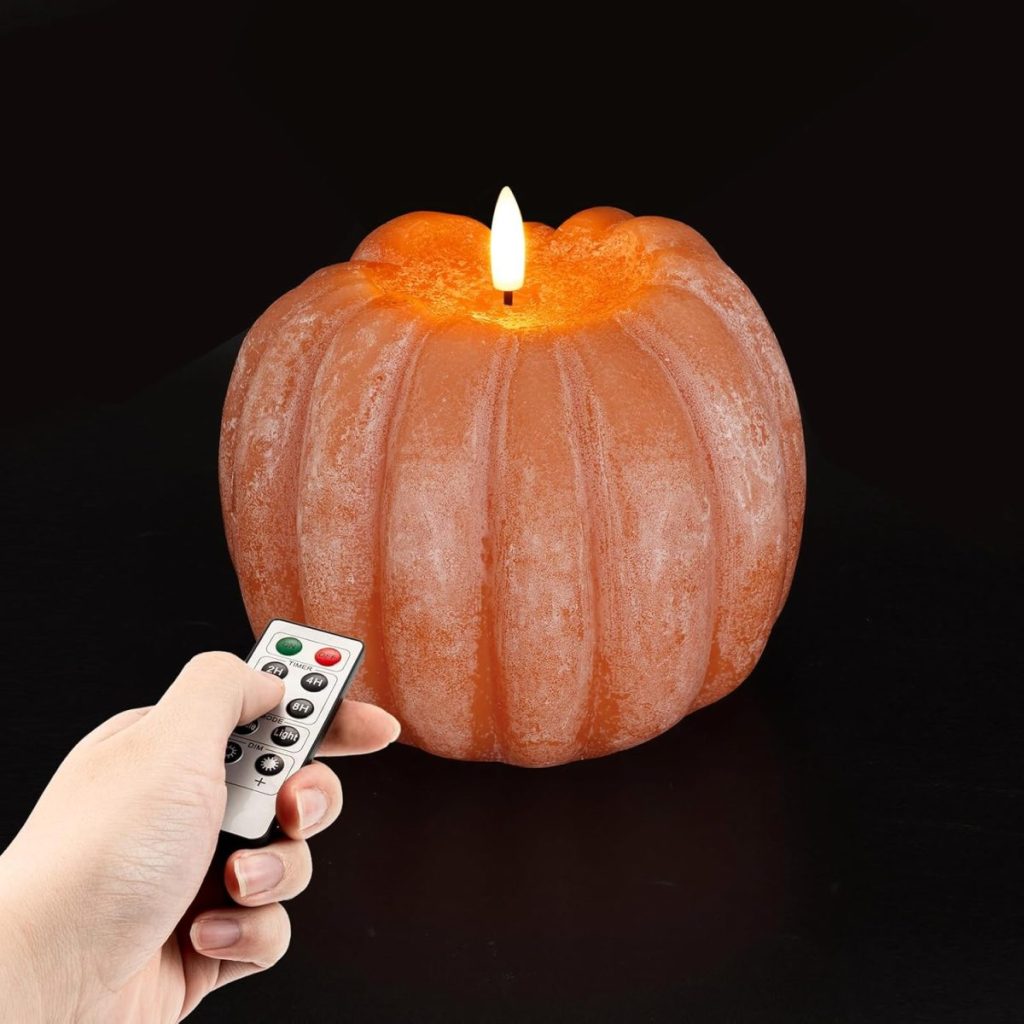 A hand holds a remote control pointed at an orange pumpkin-shaped candle with a lit flame, set against a black background.