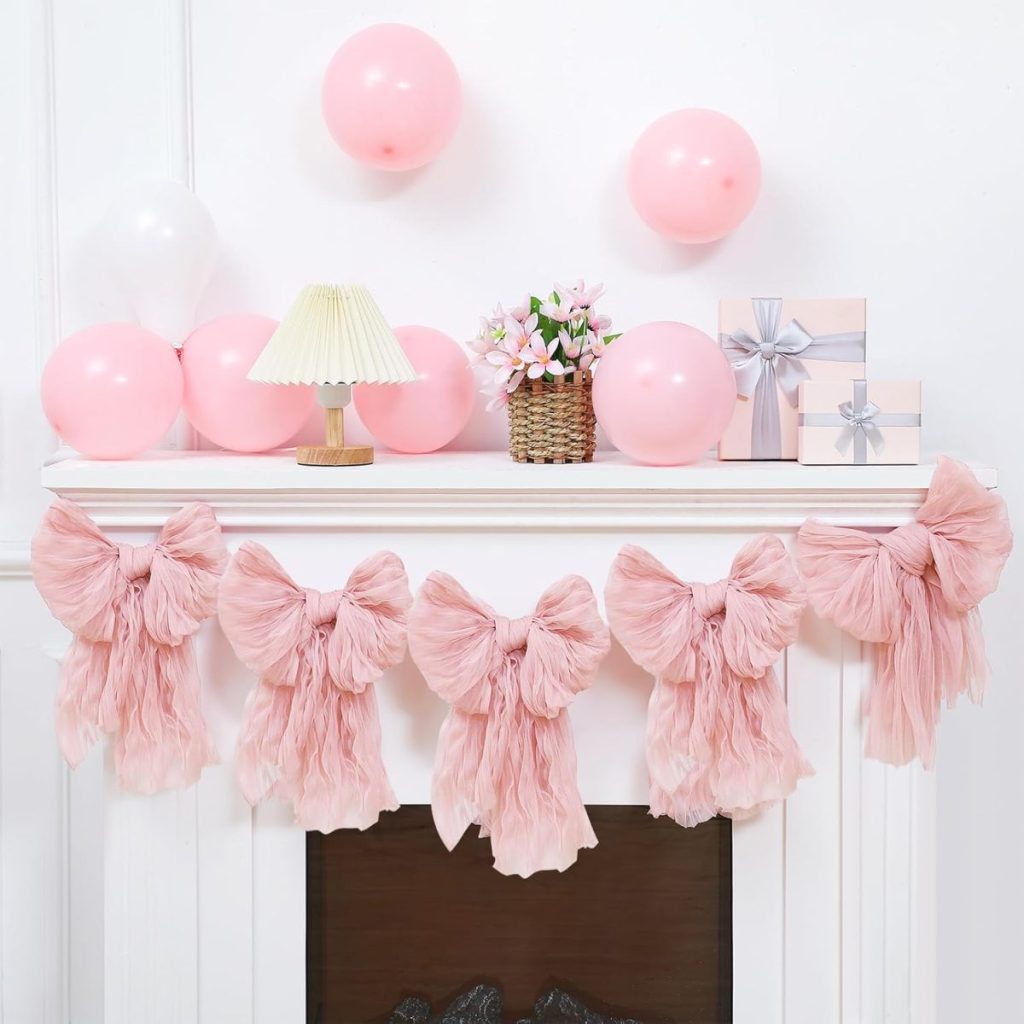 A white mantel decorated with pink fabric bows, pink and white balloons, gift boxes with silver ribbons, a small lamp, and a flower arrangement in a basket.