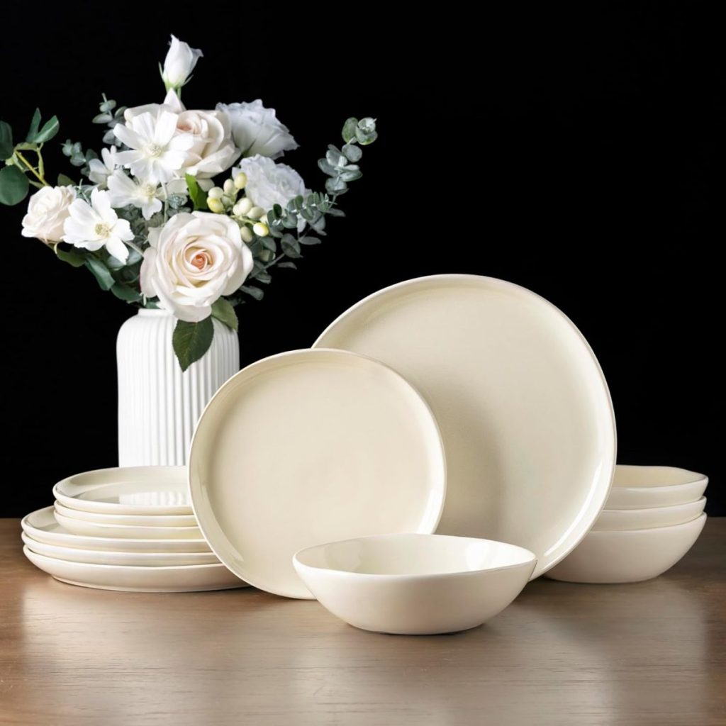 A set of cream-colored handmade clay ceramic plates and bowls arranged on a table, with a white vase of white and pale pink flowers in the background.