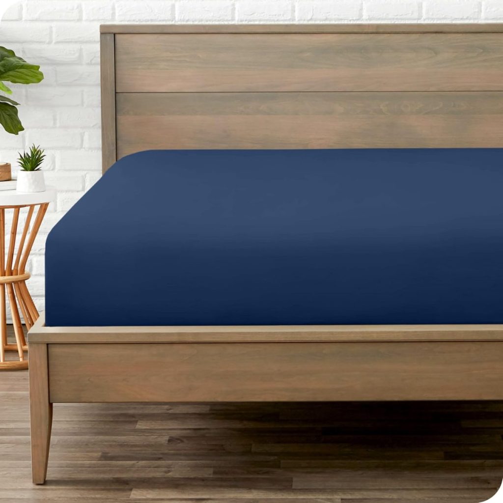A wooden bed frame with a navy blue fitted sheet in a bedroom with white brick walls, a small plant, a wooden chair, and wood flooring.
